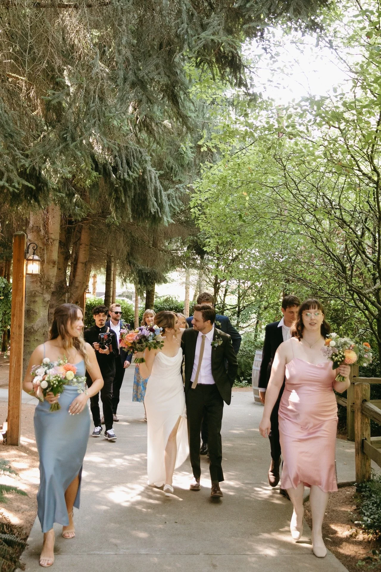 Bride, groom, and wedding party laugh and walk together down a shaded garden path after the ceremony at their garden party wedding.