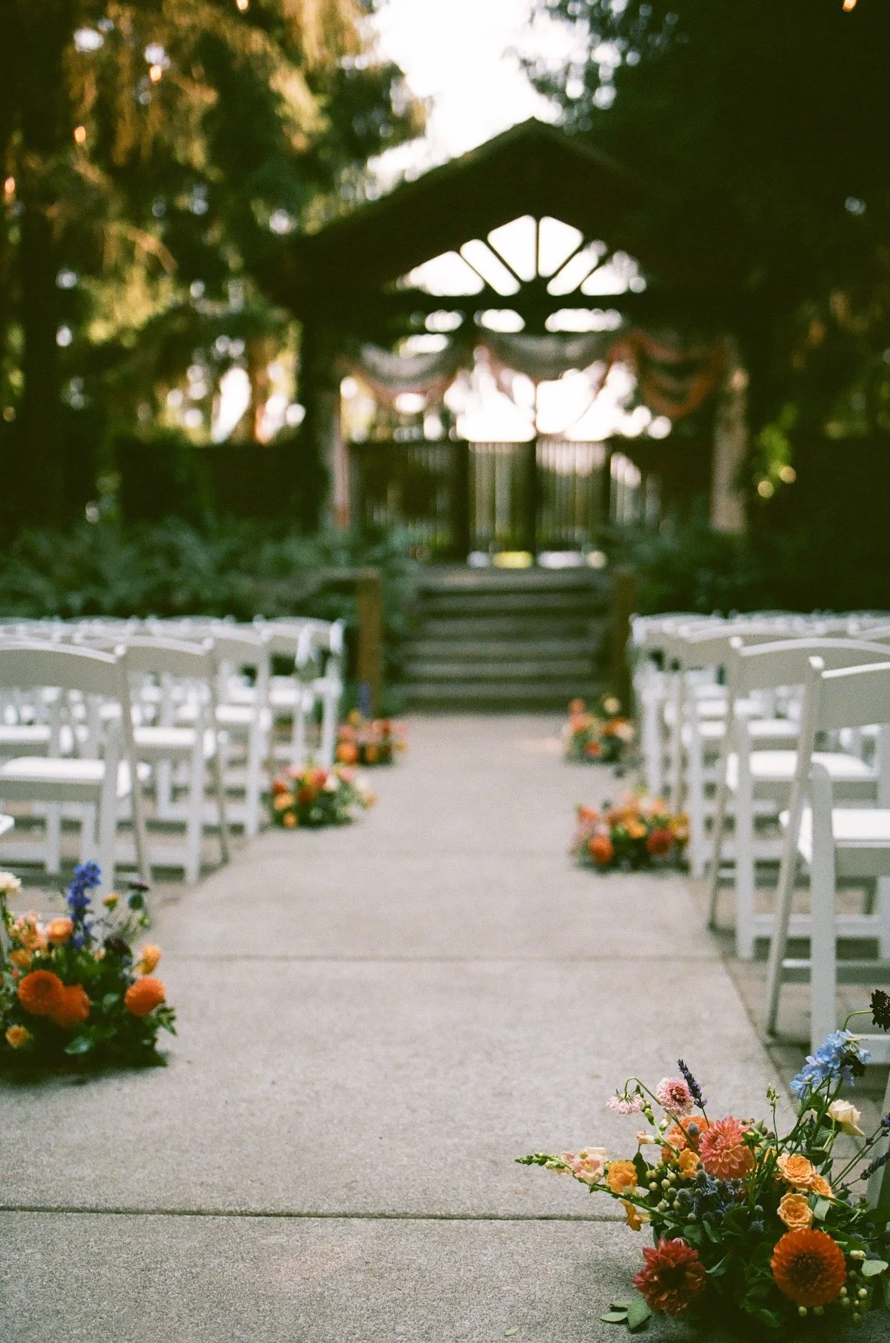 Ceremony aisle lined with white chairs and floral arrangements leading to a wooden arbor draped in fabric.