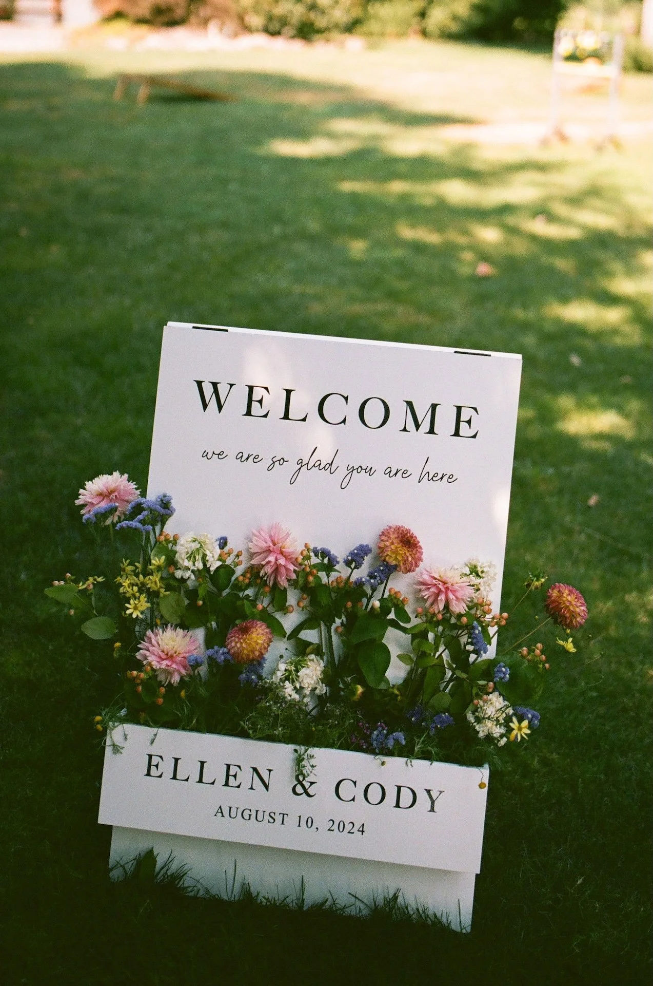 Welcome sign decorated with fresh flowers reading “Ellen &amp; Cody, August 10, 2024” greets guests at the garden party wedding.