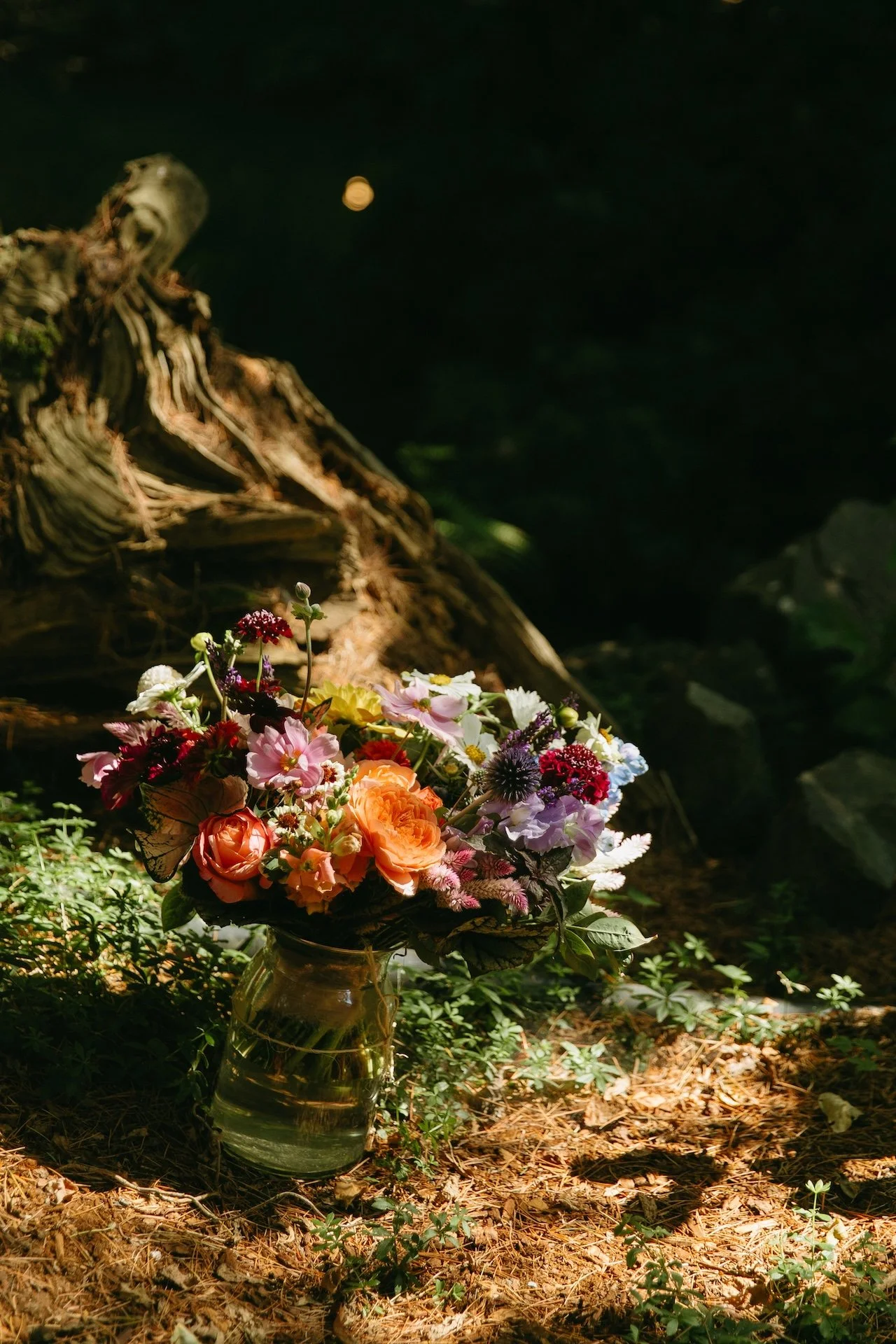 Vibrant bouquet of orange, pink, and purple flowers rests in a glass vase on the ground during a garden party wedding.