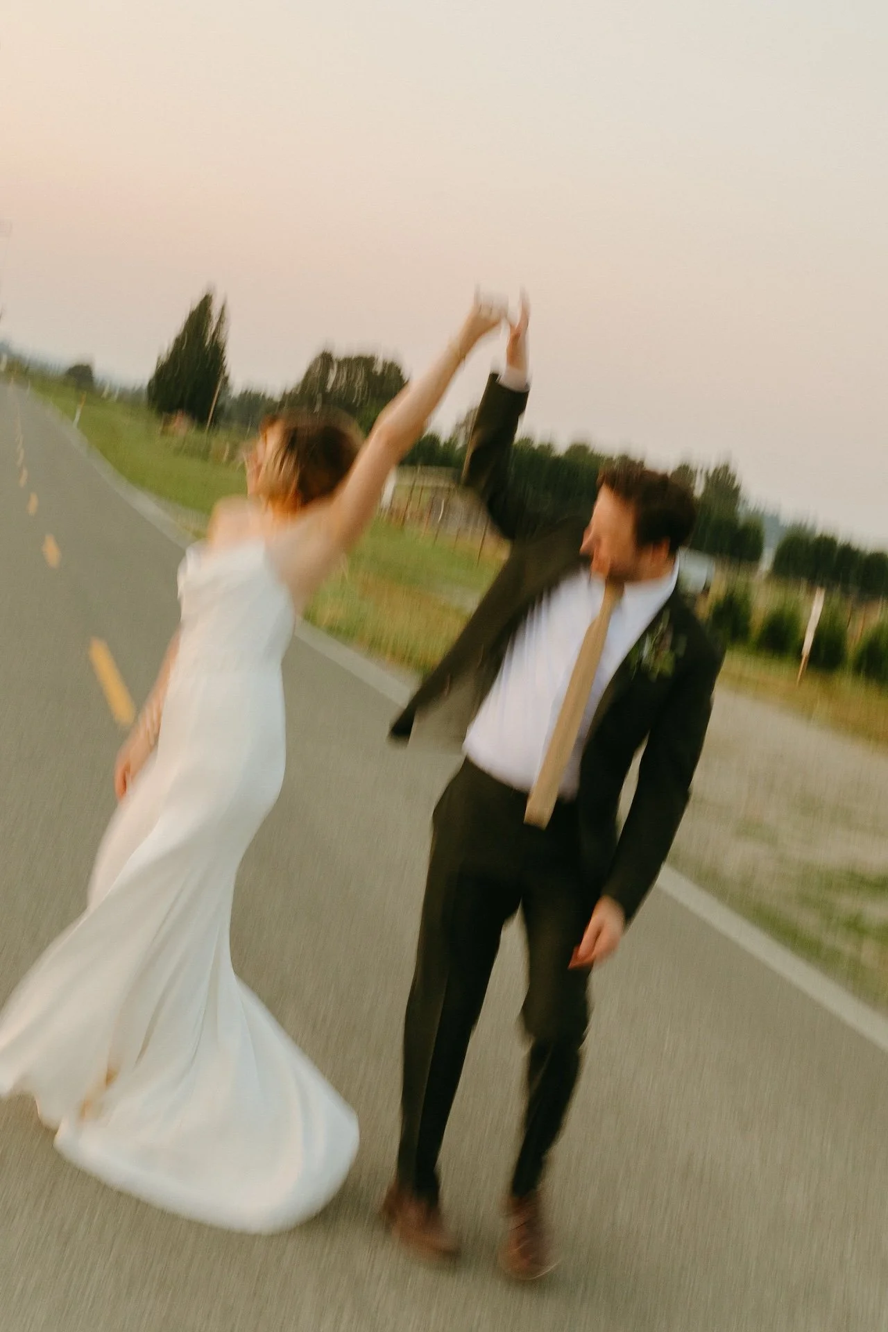 Groom twirls the bride in her satin gown on a quiet country road, both laughing in the evening light.