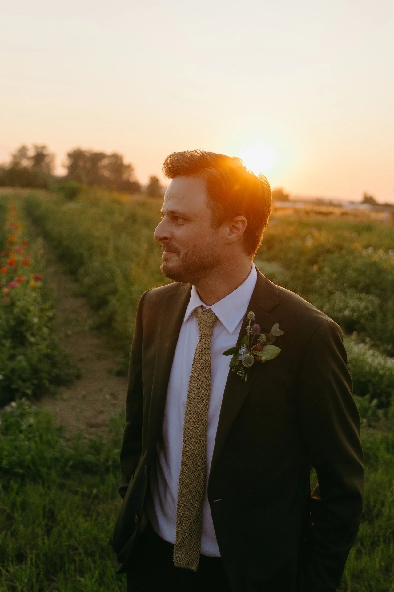 Groom stands in the field with the sun setting behind him, smiling during his garden party wedding portraits.