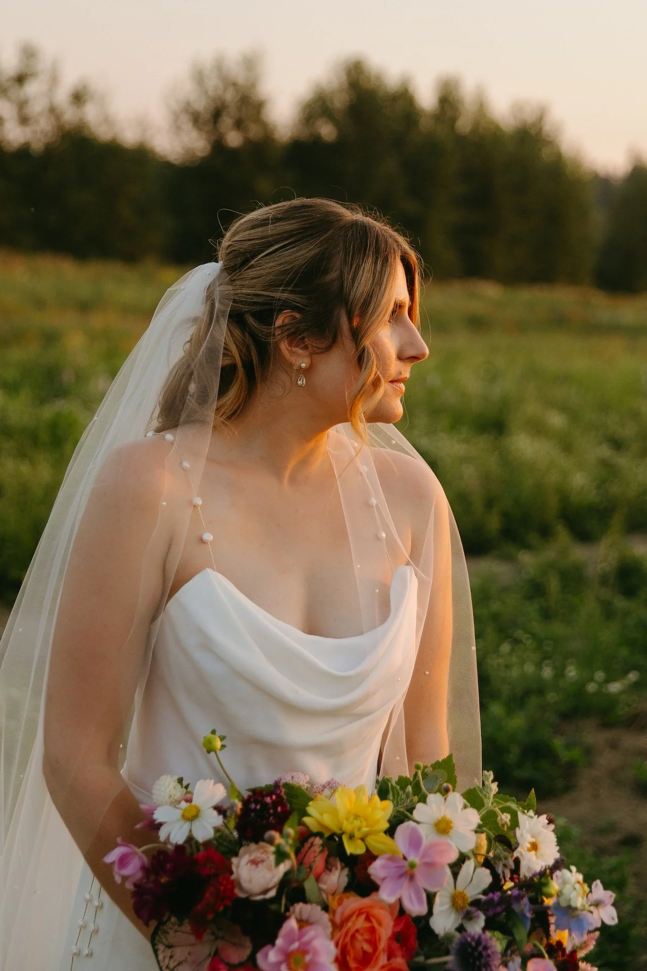 Bride holds a colorful bouquet of wildflowers and roses, her veil dotted with pearls in the evening light.