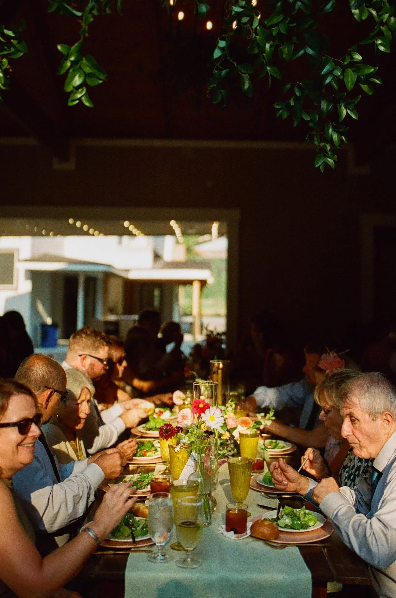 Guests sit at long tables enjoying dinner, the table lined with colorful flowers and candles at the garden party wedding reception.