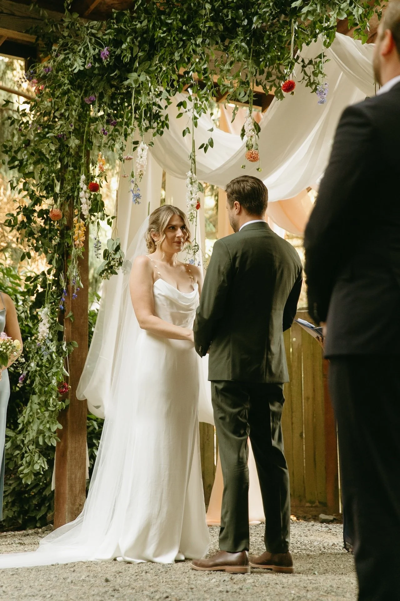 Bride and groom exchange vows under a floral-draped arbor, framed by greenery and draped fabric.