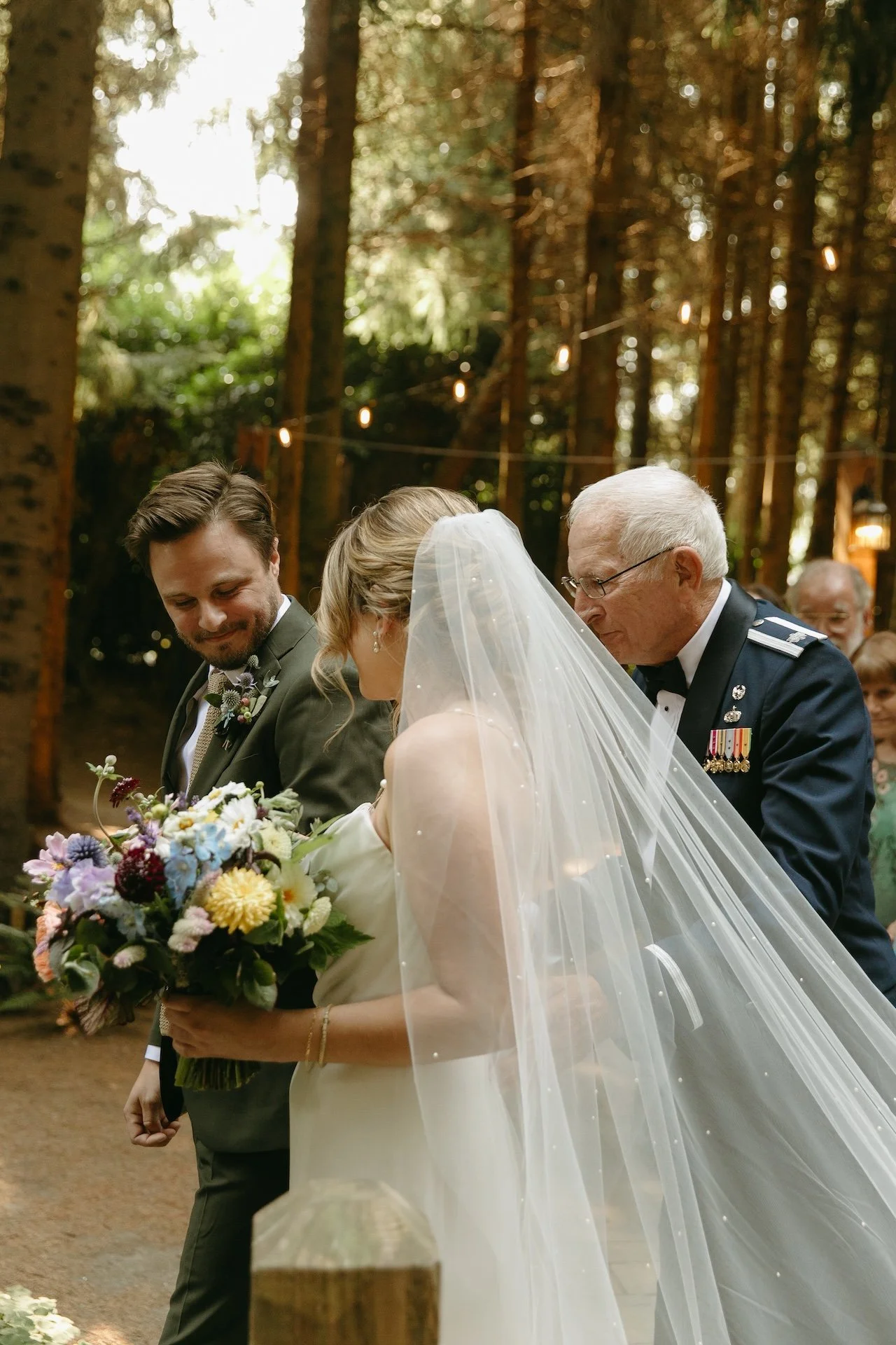 Bride walks down the aisle with her father, holding a colorful bouquet, while the groom smiles at their garden party wedding.