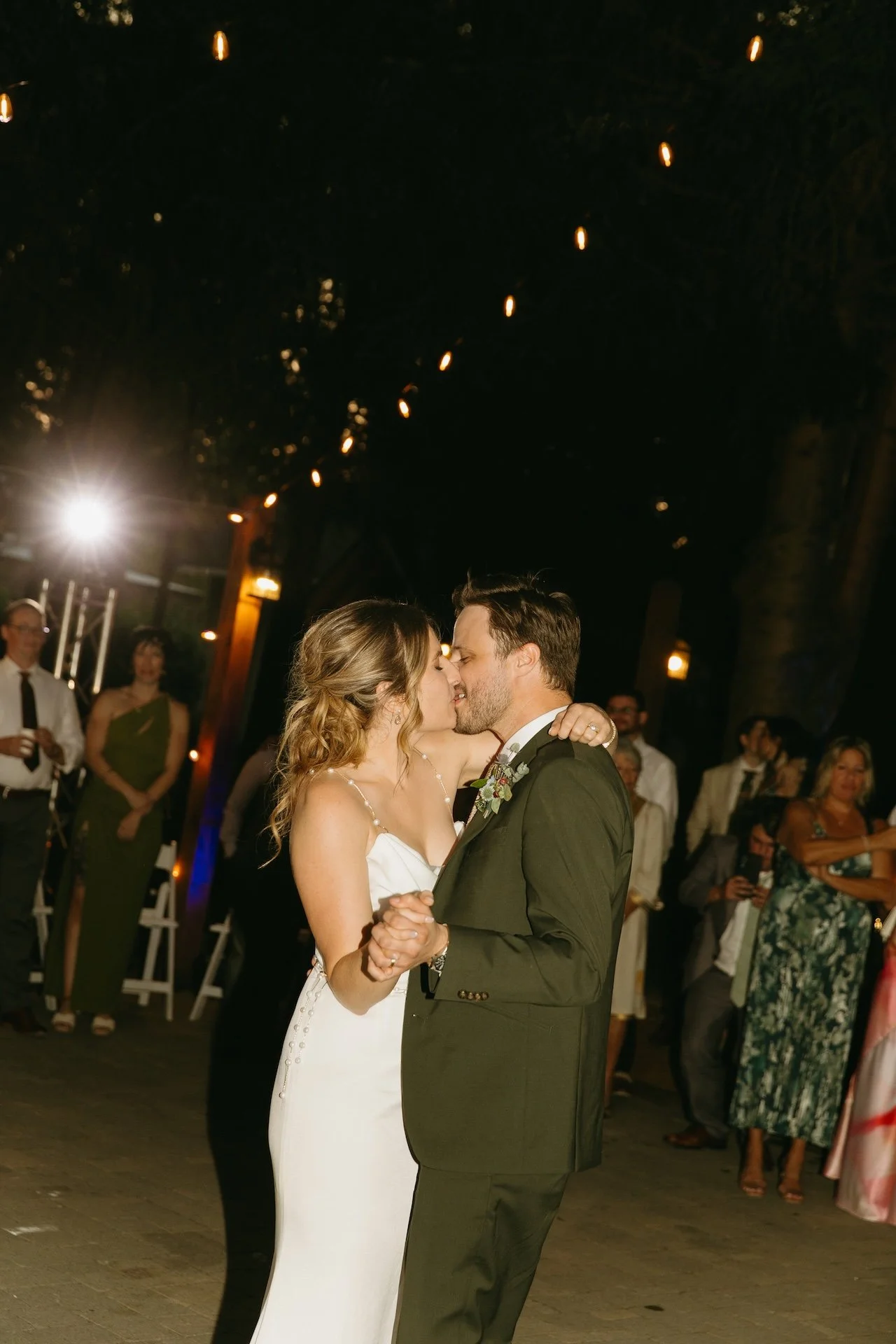 Couple kiss during their first dance beneath glowing string lights at their garden party wedding.
