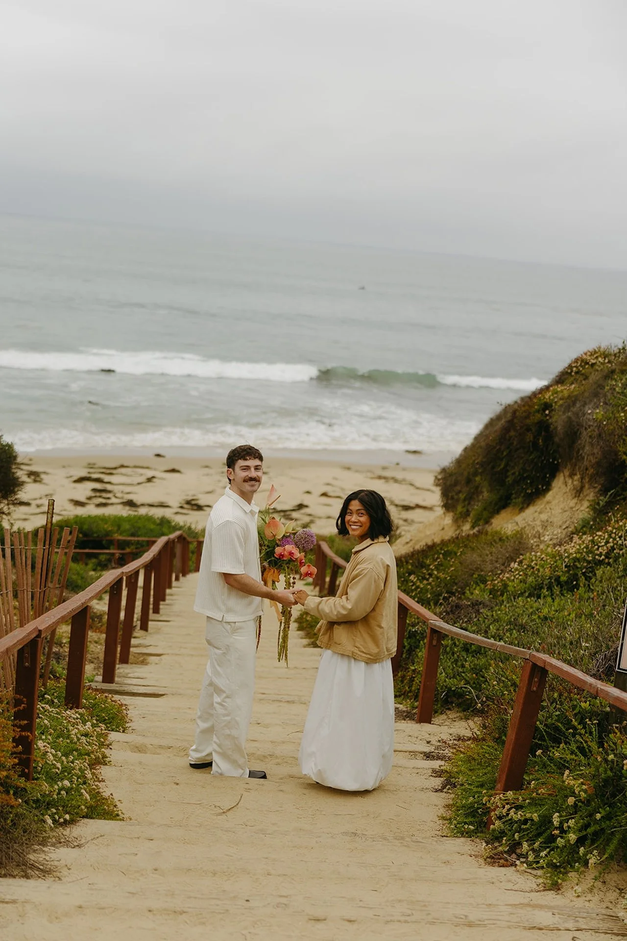 The bride and groom turning around to smile at the camera as they hold hands and walk down to the beach for their elopement in laguna beach