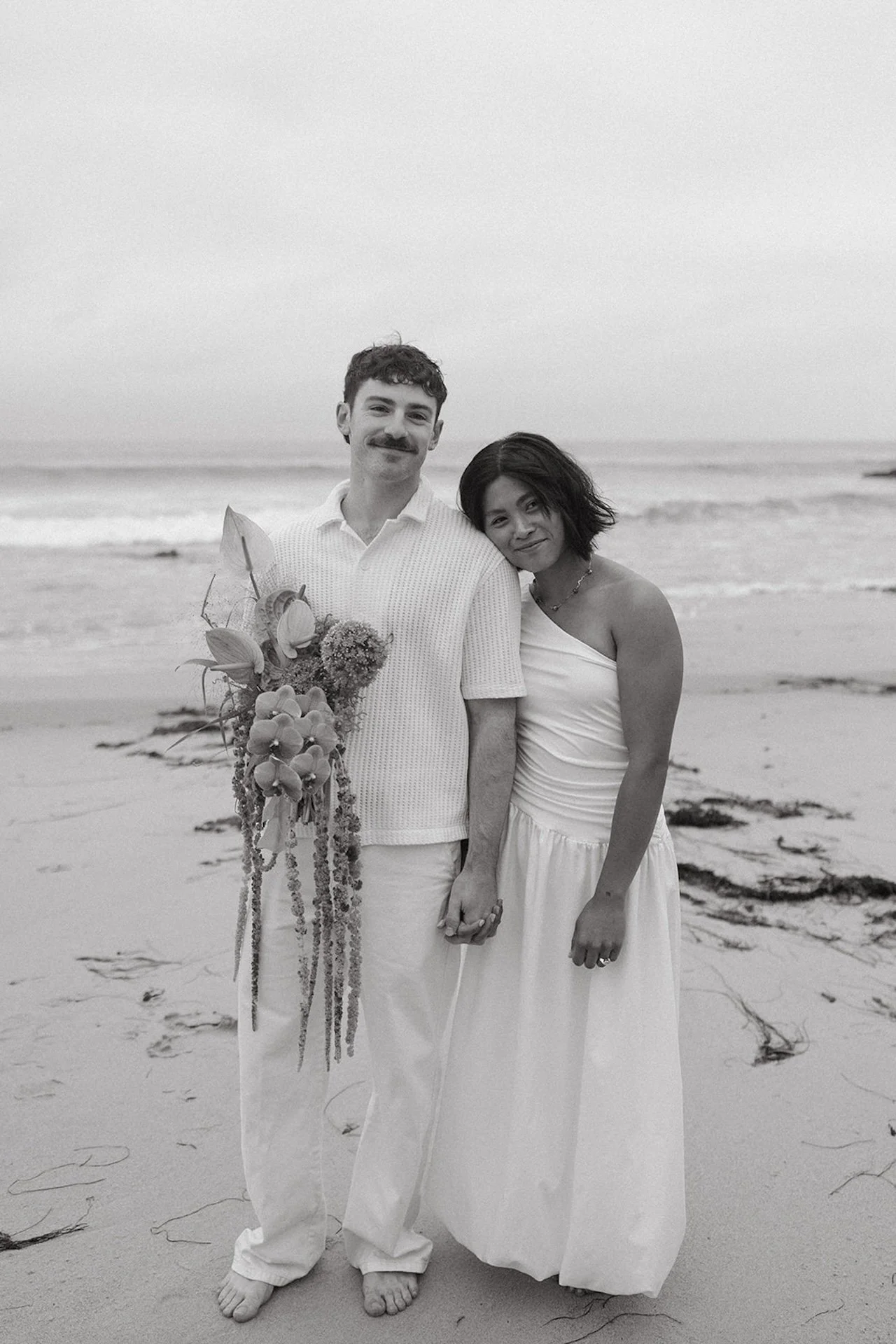 The bride and groom standing on the beach holding hands as she leans her head on his shoulder.
