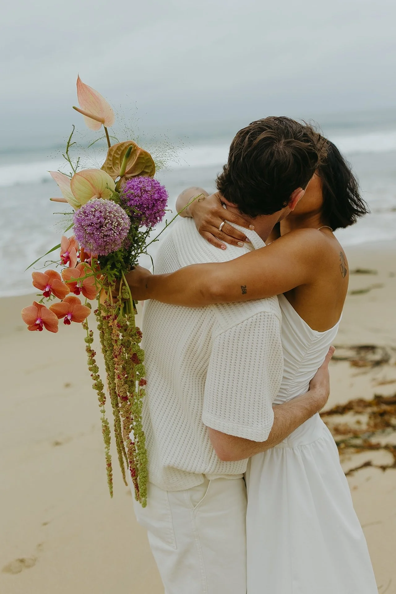 The bride and groom kissing on the beach while she holds her bridal bouquet over his shoulder during their elopement on the beach