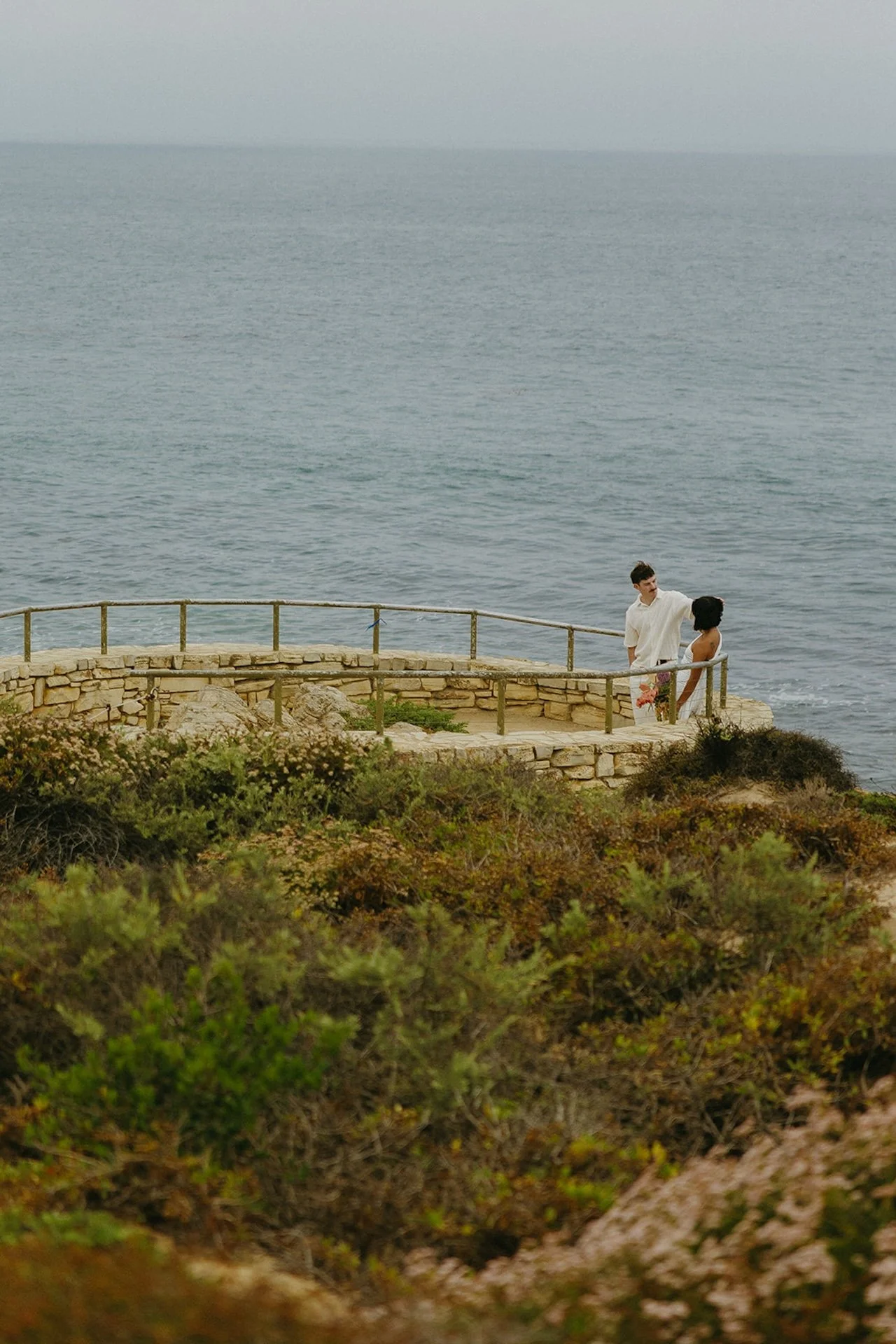 A far away shot of the bride and groom standing at their ceremony spot that overlooks the beach during their beach elopement