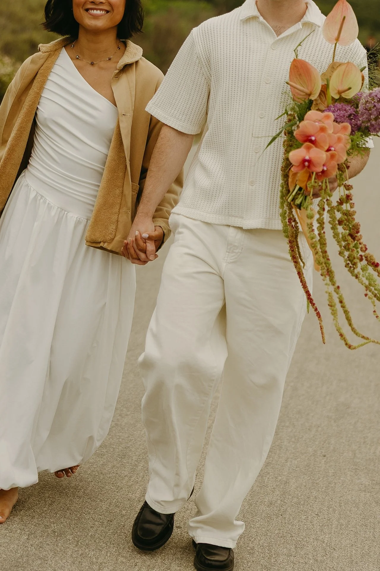 A detail shot from the shoudlers down of the bride and groom holding hands and walking down a path towards their elopement on the beach