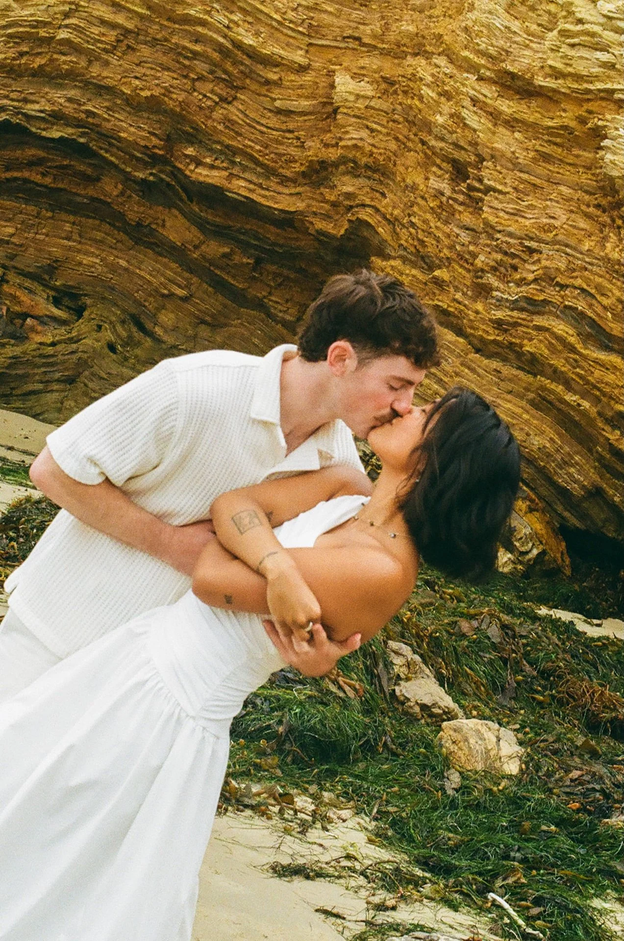 The bride and groom kissing and hugging as they take photos on the beach with a coastal cliff in the background