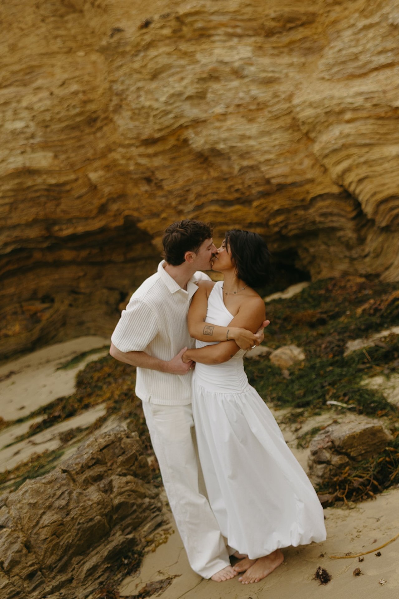 The bride and groom kissing and hugging as they take photos on the beach with a coastal cliff in the background