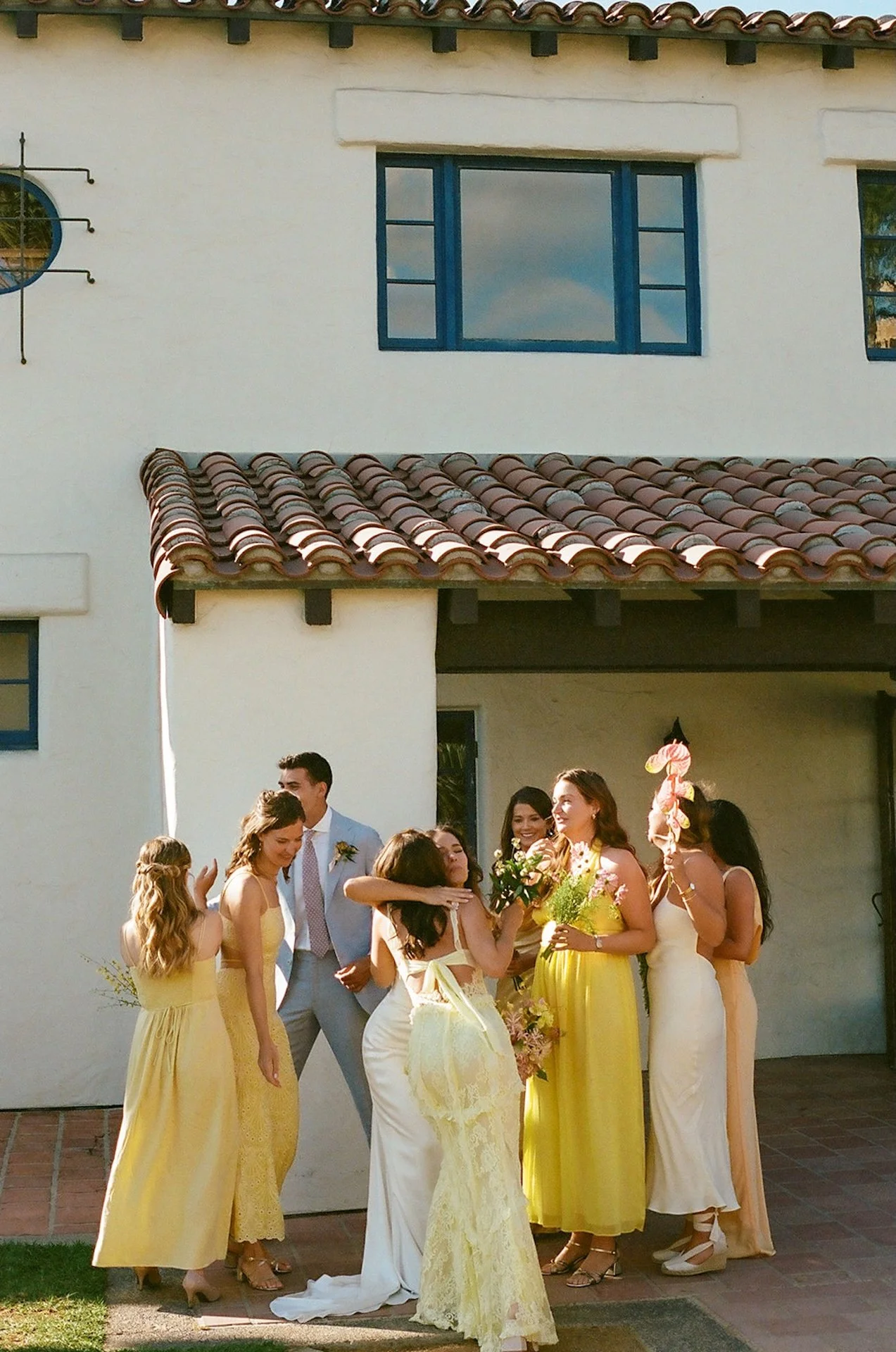 Bridesmaids in soft yellow tones surround the bride in a warm, sunlit courtyard moment.