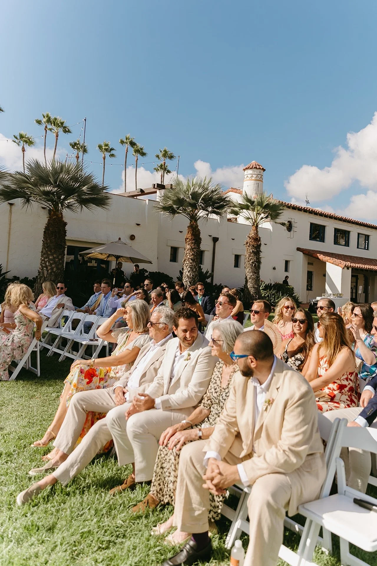 Guests sit beneath bright sunshine and palm trees at a San Juan Capistrano Wedding Venue.