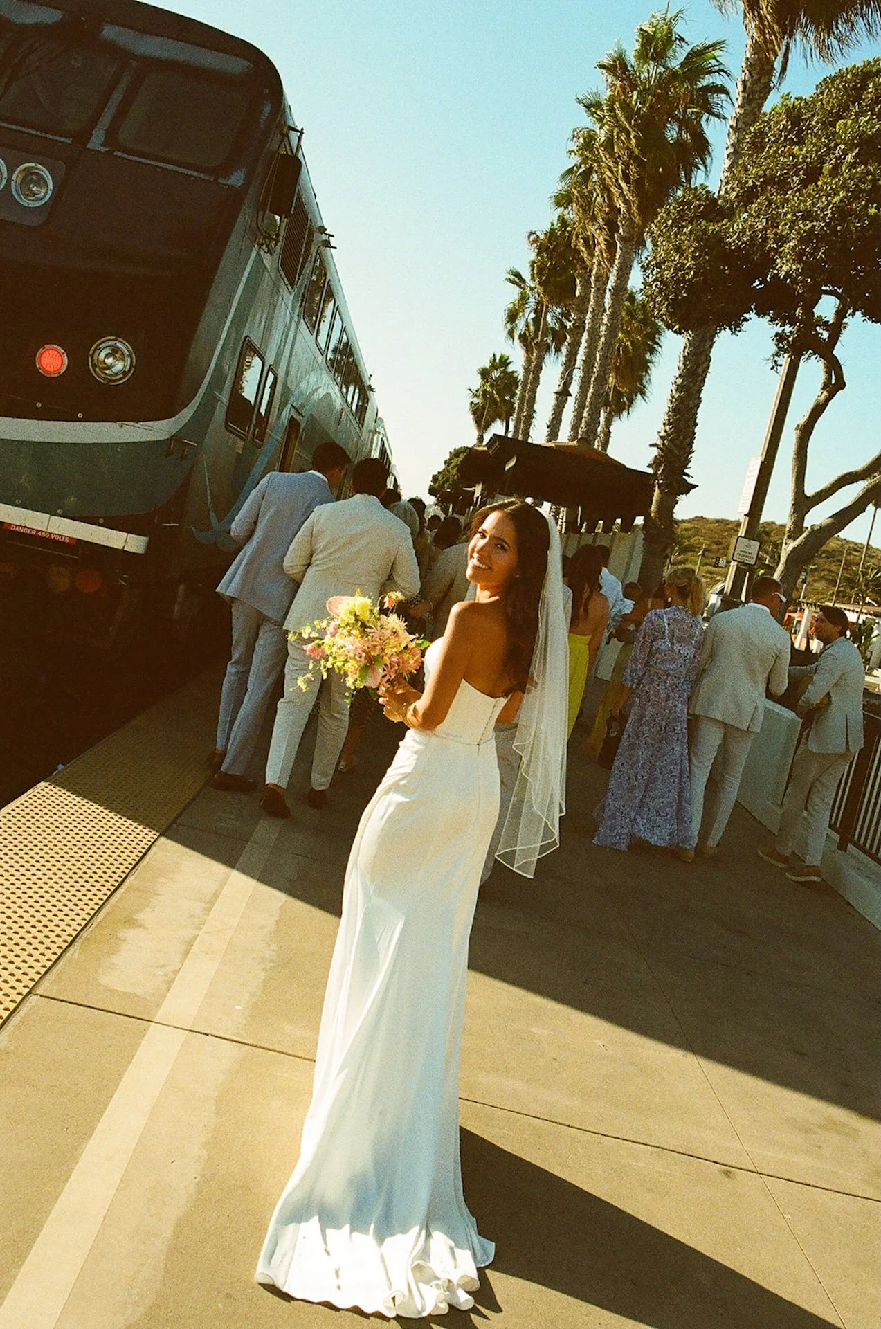 The bride looks back over her shoulder with a smile as guests board the train behind her.