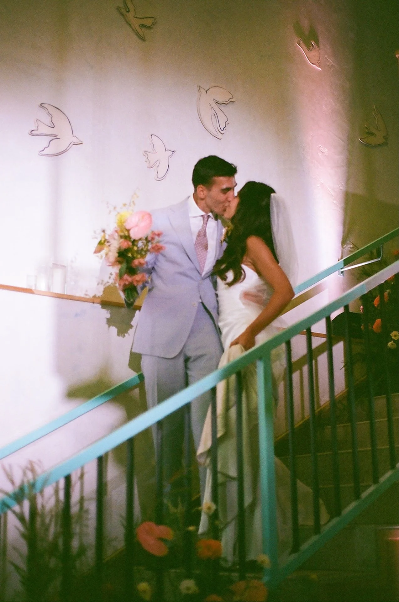 The groom holding the wedding bouquet as the bride and groom walk down the stairs and share a kiss on their way to their San Juan Capistrano Wedding venue reception