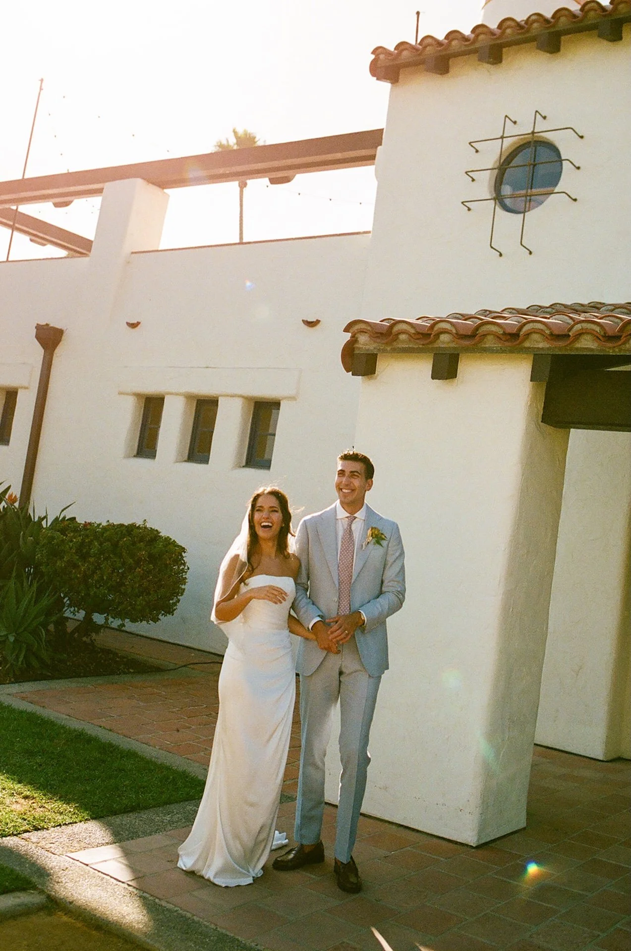 The bride and groom laughing in front of a white and orange stucko building during their bride and groom portraits