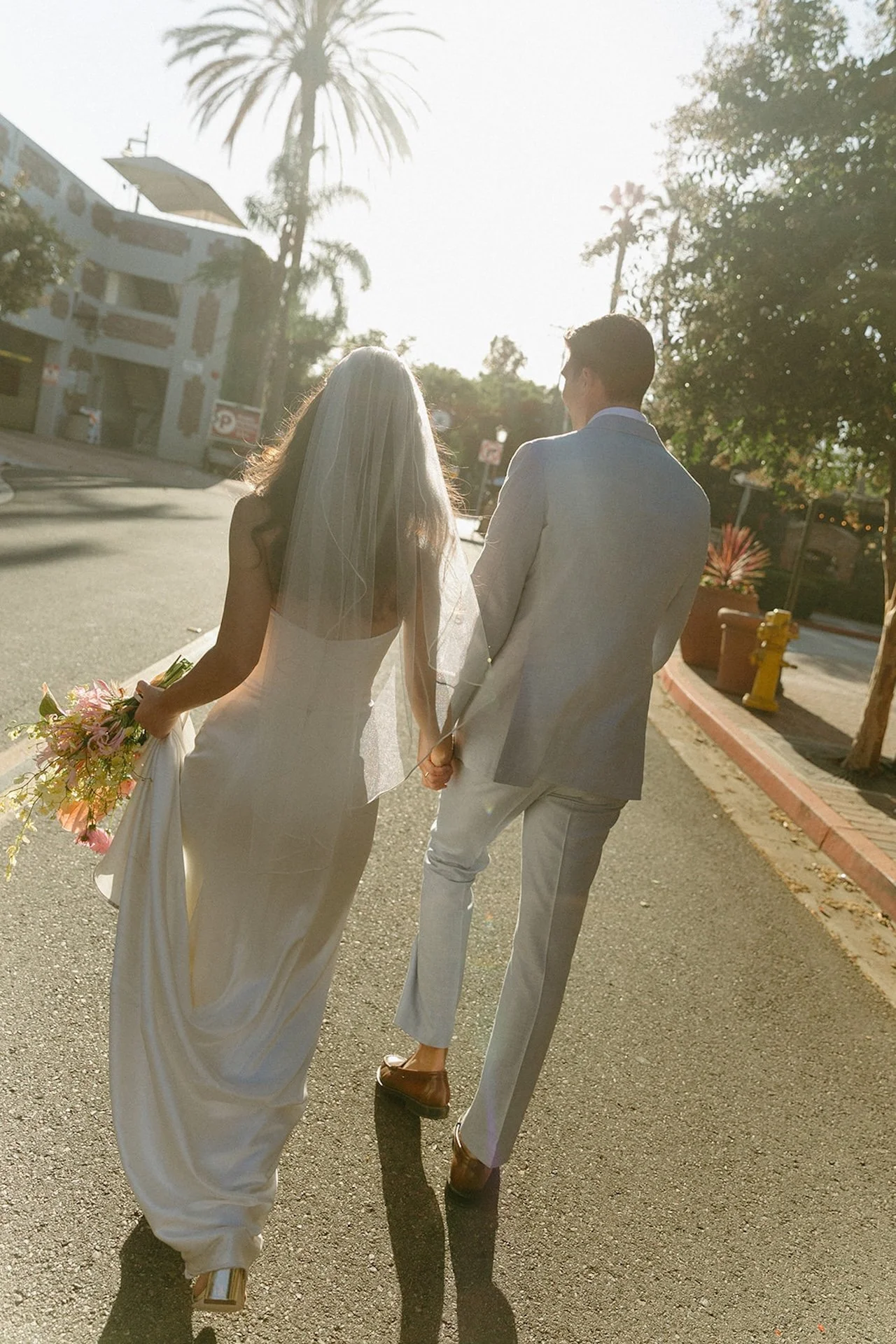 The bride and groom walking into the sunset after their wedding as the bride holds her wedding bouquet and dress in her hands.
