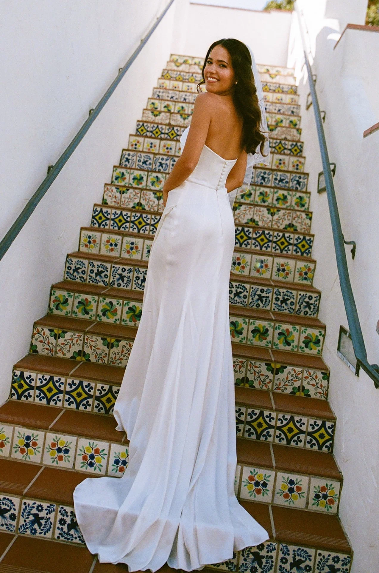 The bride turned around and looking at the camera with a soft smile on a set of spanish tile stairs at their San Juan Capistrano Wedding venue