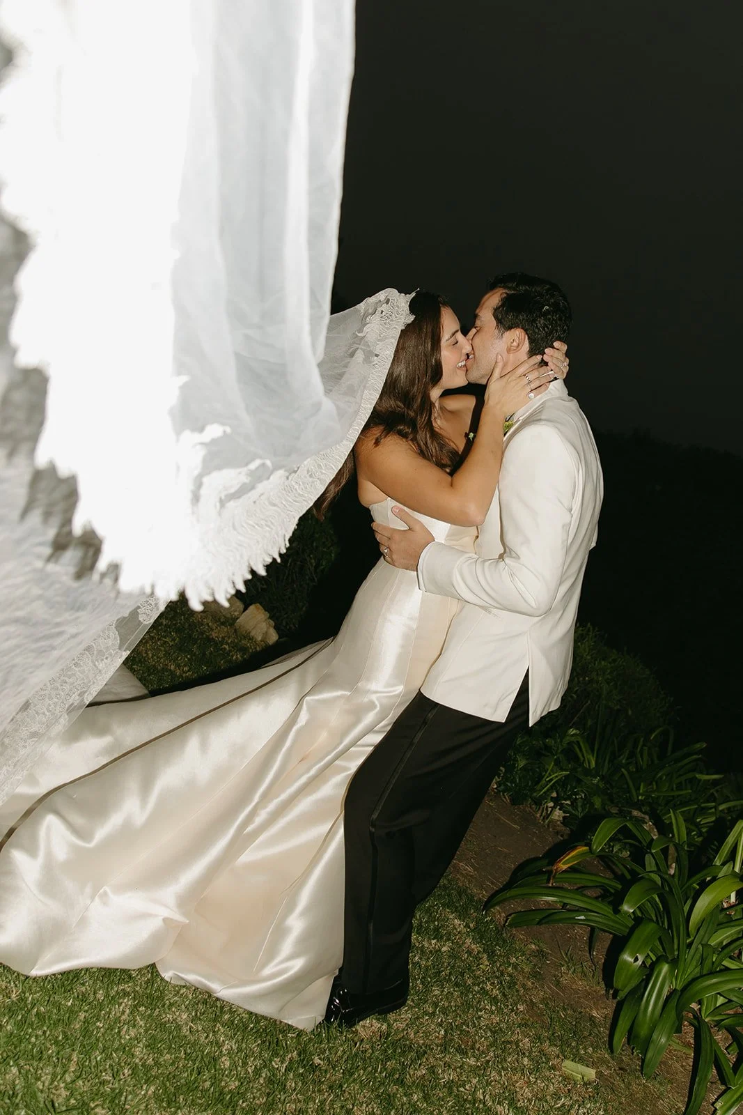 Bride and groom sharing a quiet kiss at night, her veil blowing dramatically in the wind at a Palos Verdes wedding venue.