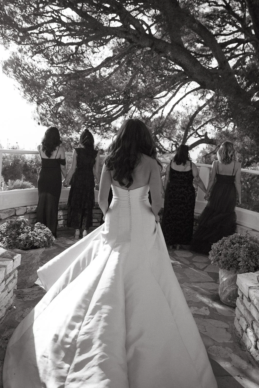 Black-and-white image of the bride walking forward as bridesmaids stand hand in hand ahead of her.