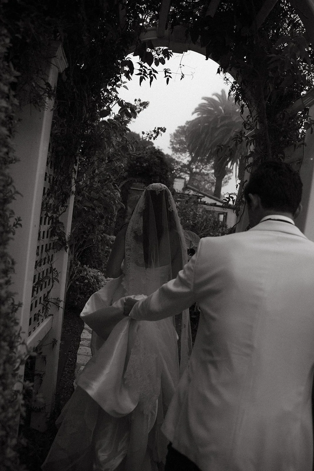 Black-and-white image of the couple walking away beneath an archway, the groom holding the bride’s train as they exit the venue.
