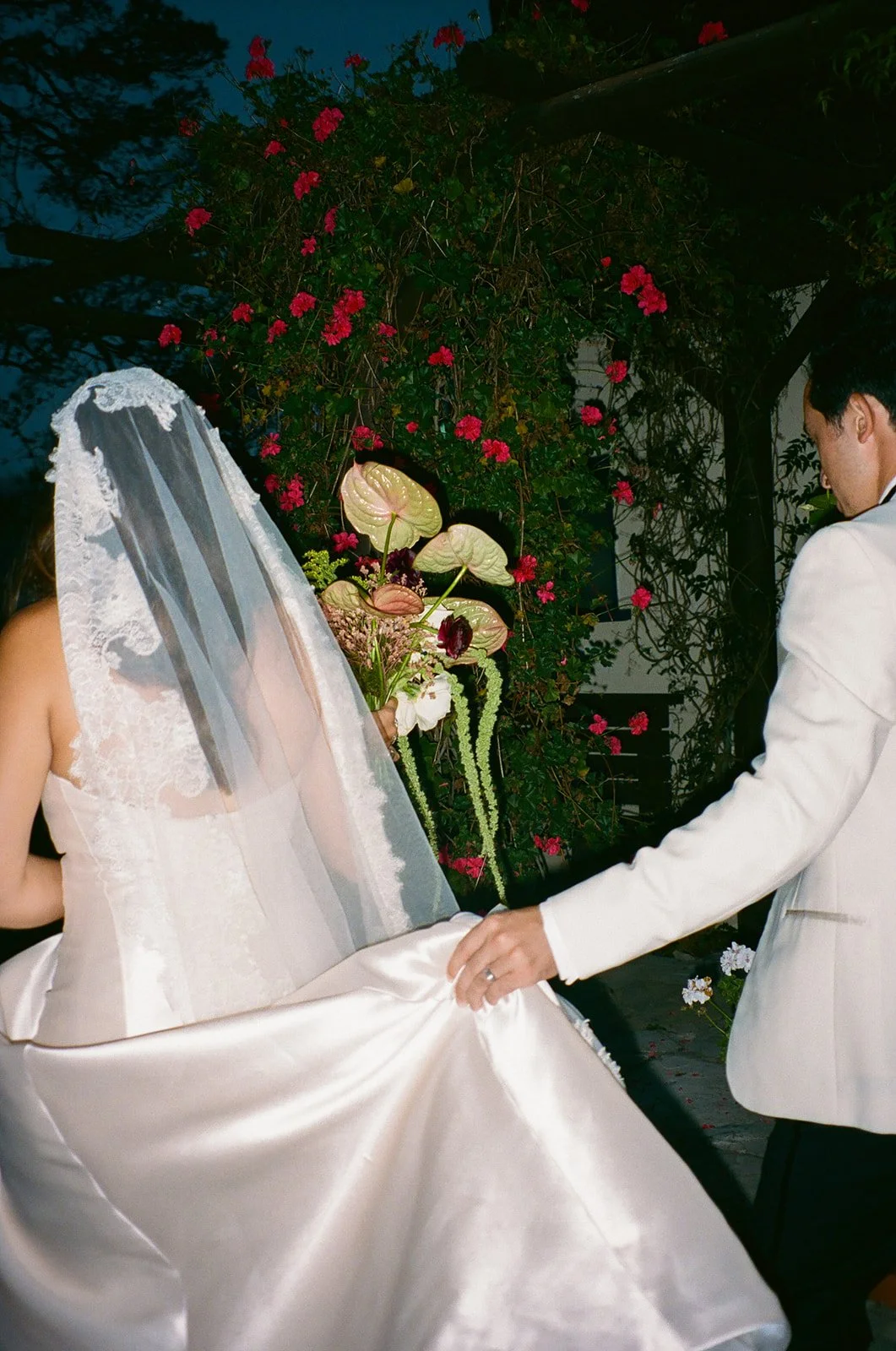 Groom gently lifting the bride’s dress as they walk together through the garden path.