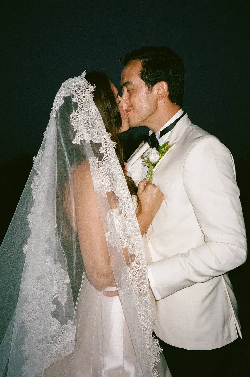 Bride and groom laughing and sharing a quiet kiss at night as her veil flows behind them at a Historic Wedding Venue.
