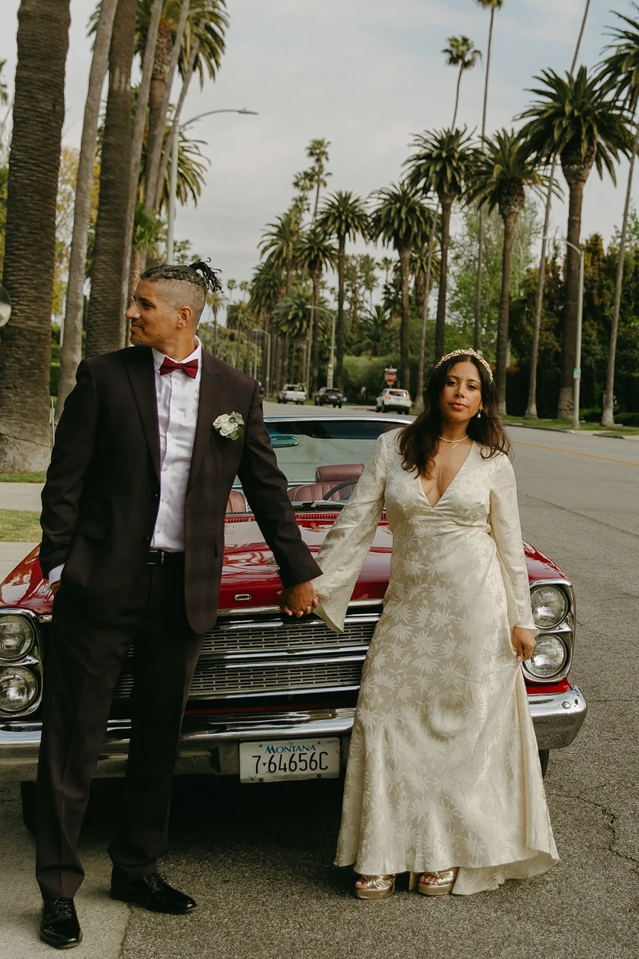 Newlyweds holding hands in an editorial pose in front of a red vintage car on a palm tree lined road in for their Beverly Hills Courthouse Elopement