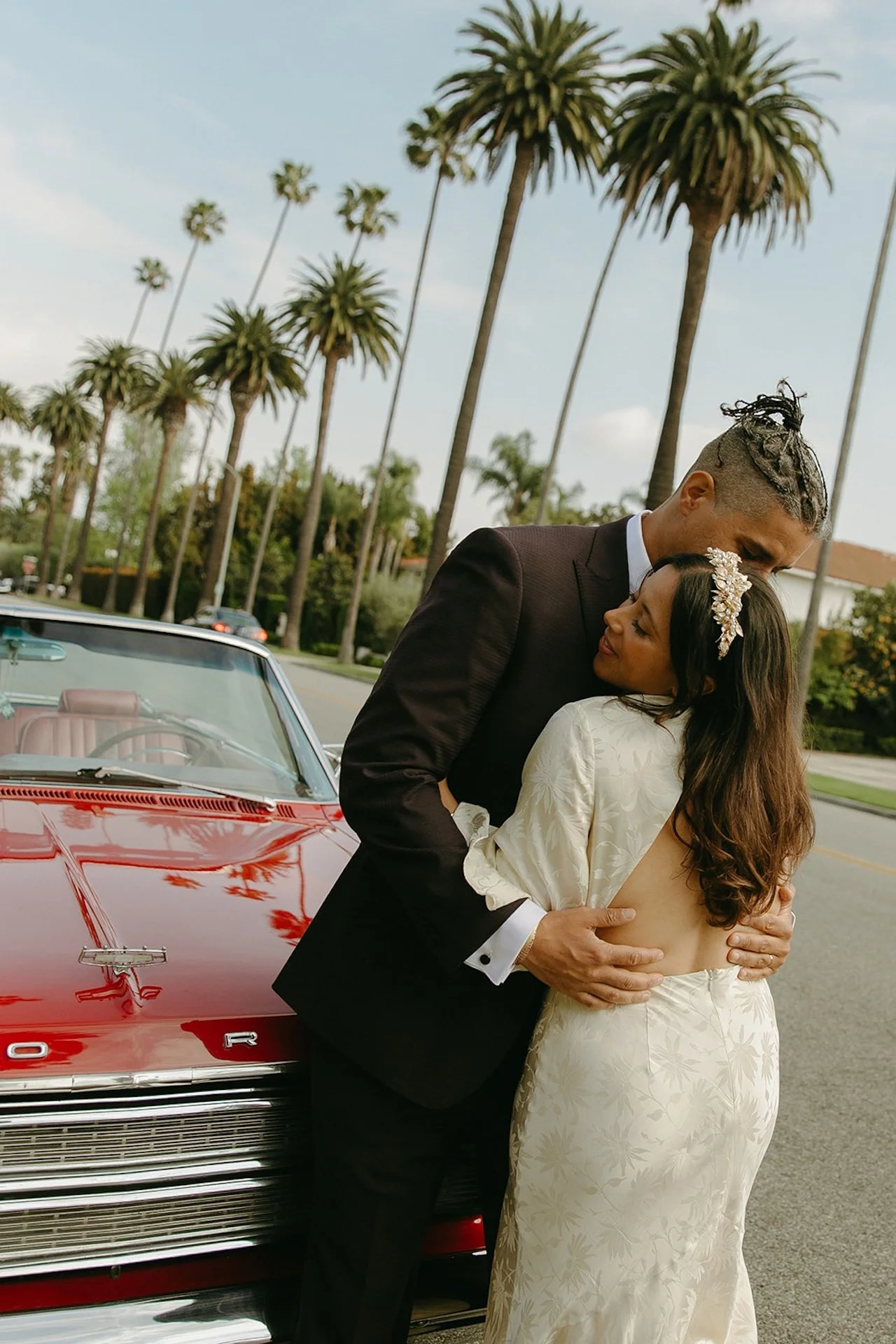Newlyweds hugging while leaning on a red convertable with palm trees lining the road in the background.