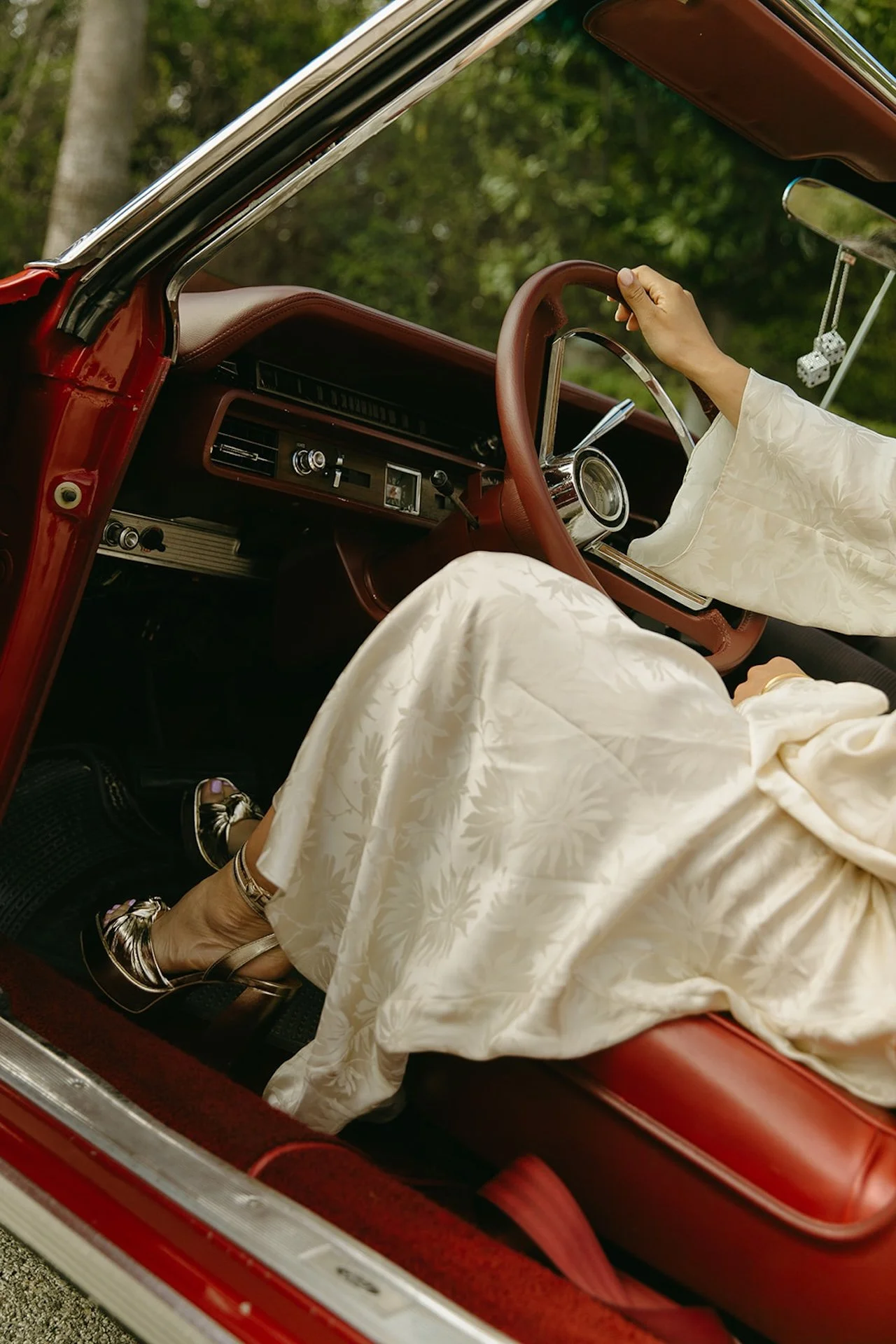 Detail photo of the bride sitting in the drivers seat of a red convertable. Showing off her silver heels, silk dress, and the red vintage convertable.