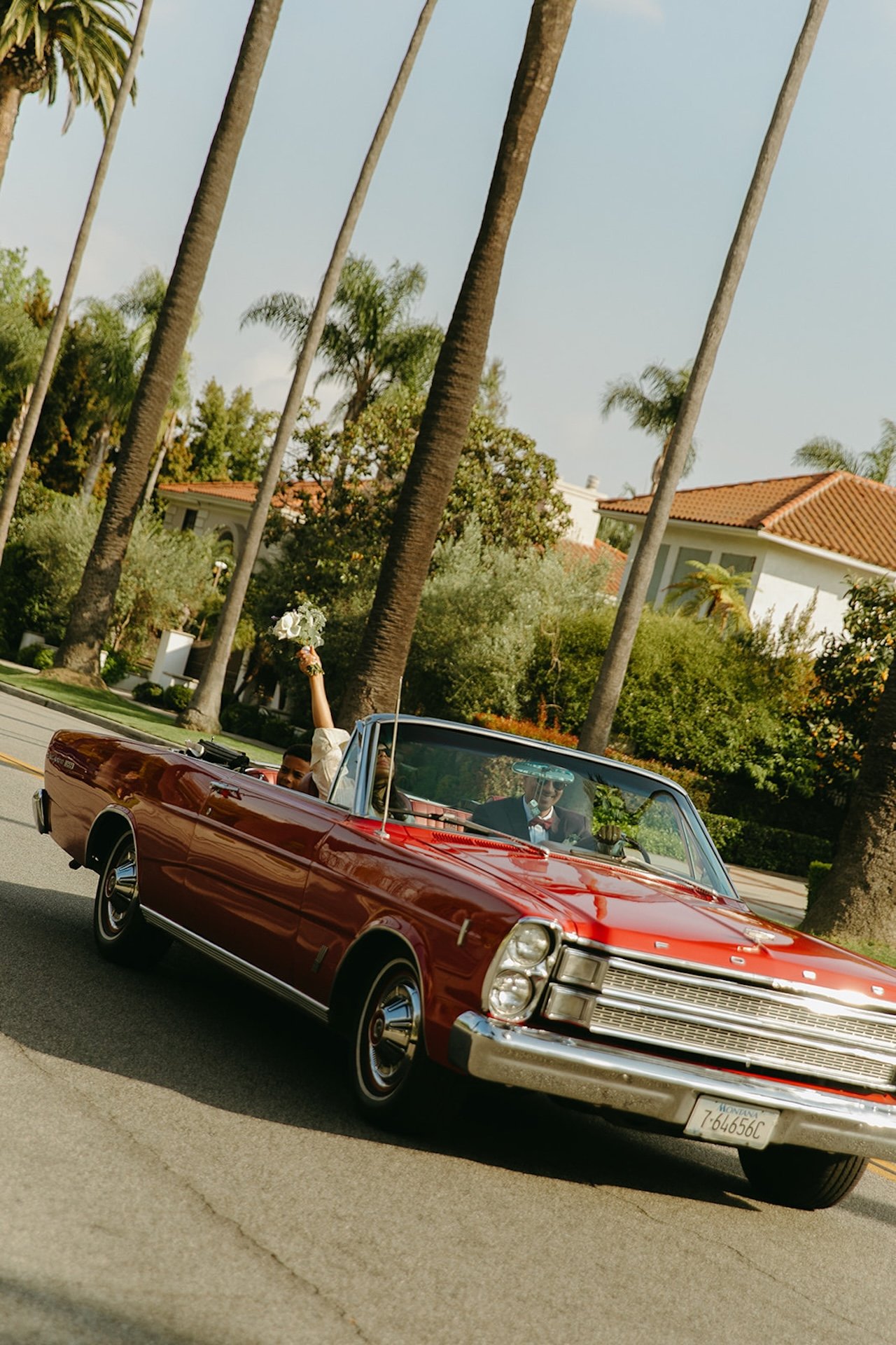 Newlyweds driving down a palm tree lined street in a red convertable after their Beverly Hills Courthouse Elopement with the bride waving her bridal bouquet in the air