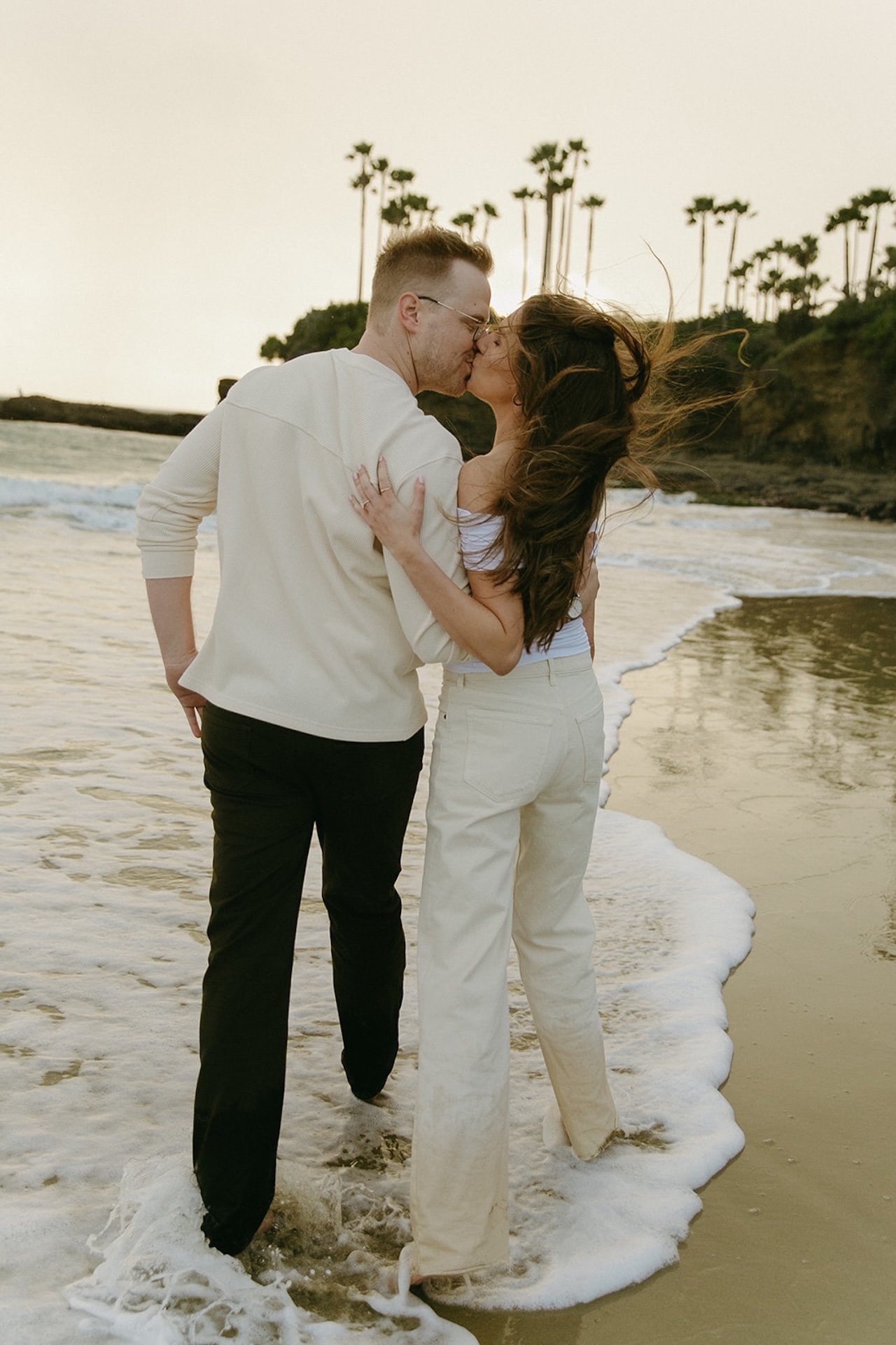 The couple kissing at the shoreline with waves washing around their feet, photographed during laguna beach engagement photos at shaws cove laguna beach.
