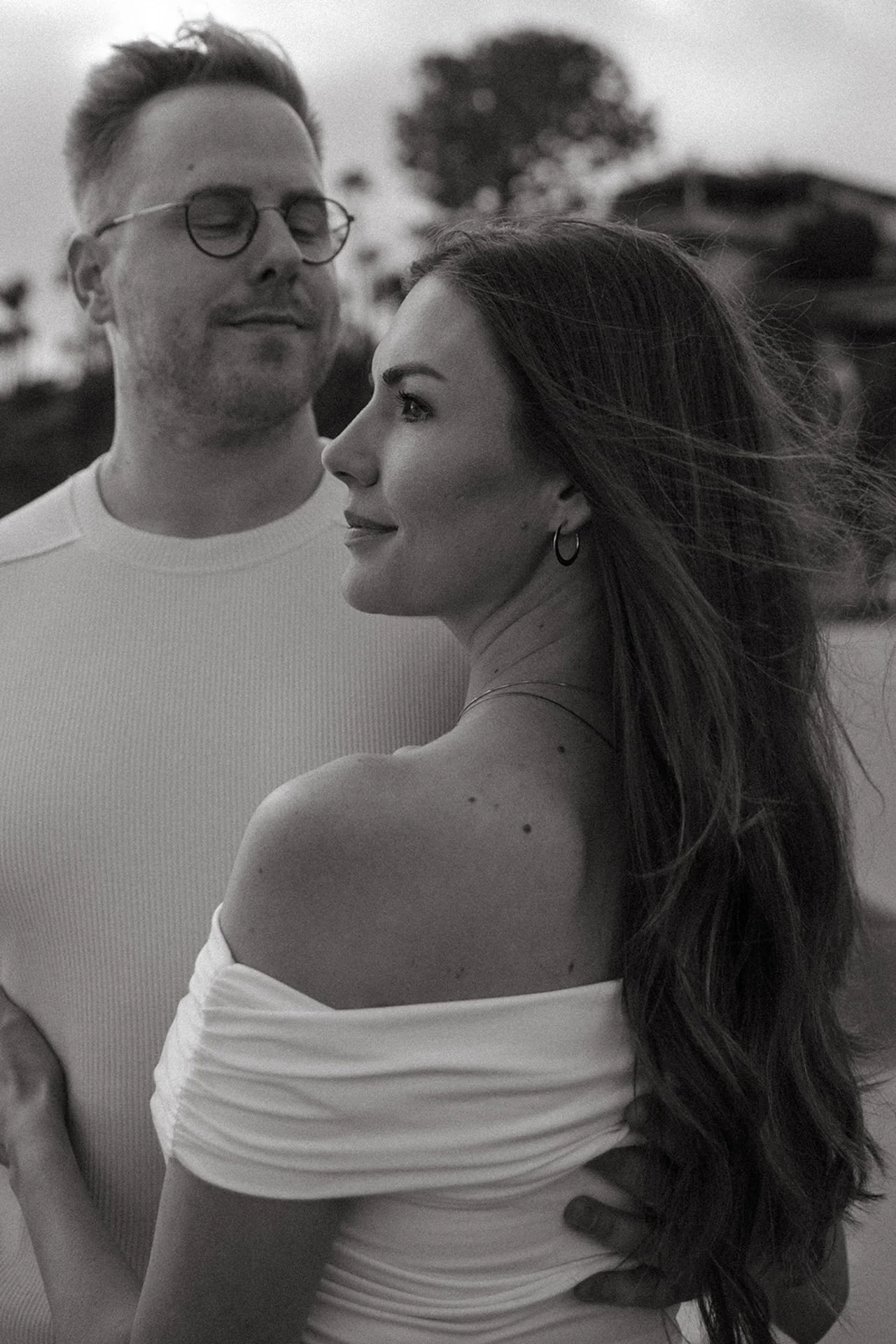 Black and white portrait of an engaged couple standing close together, wind blowing through her hair as they share a quiet moment by the beach.