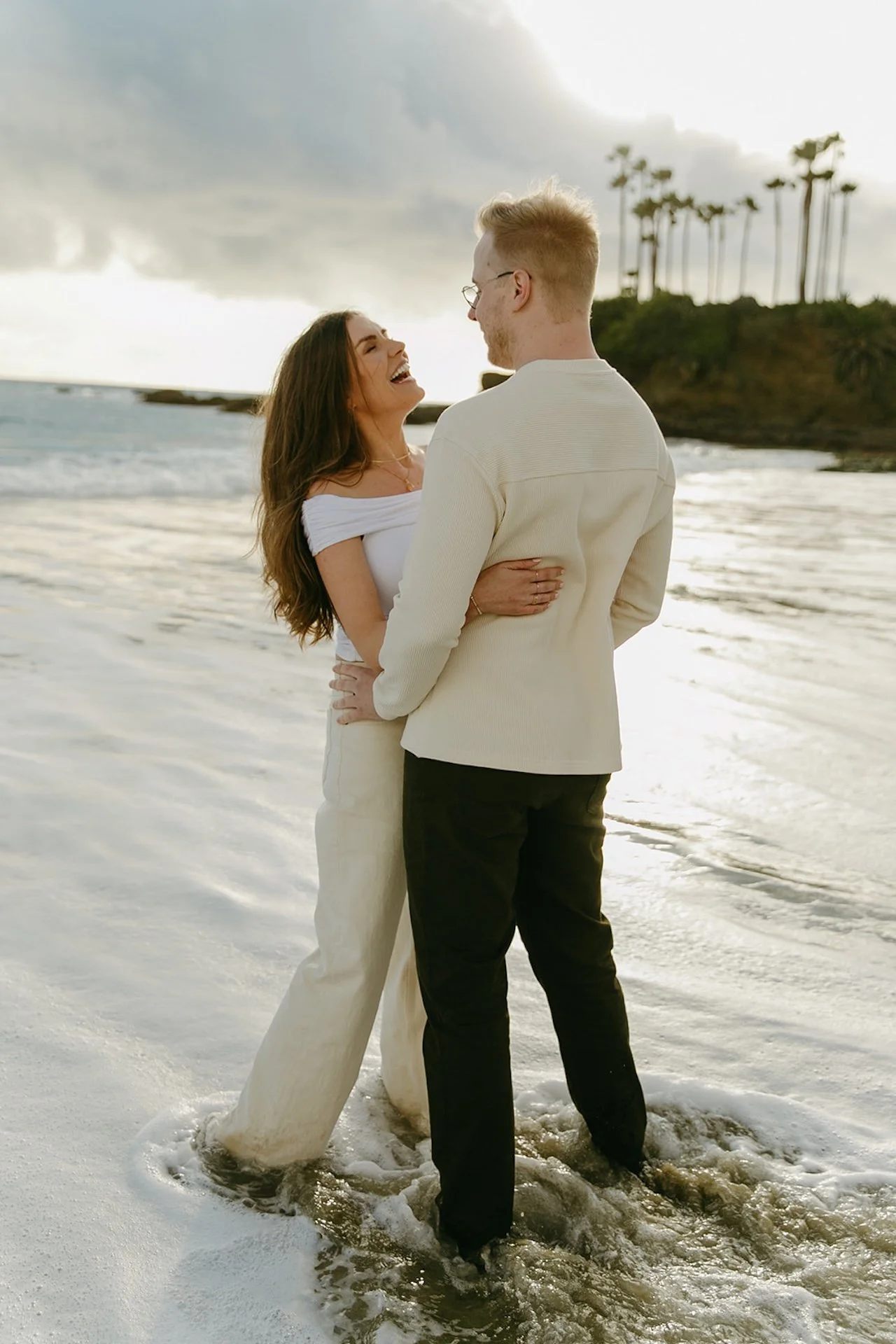 The engaged couple standing ankle-deep in the water, laughing together as the tide moves in along the sandy shoreline.