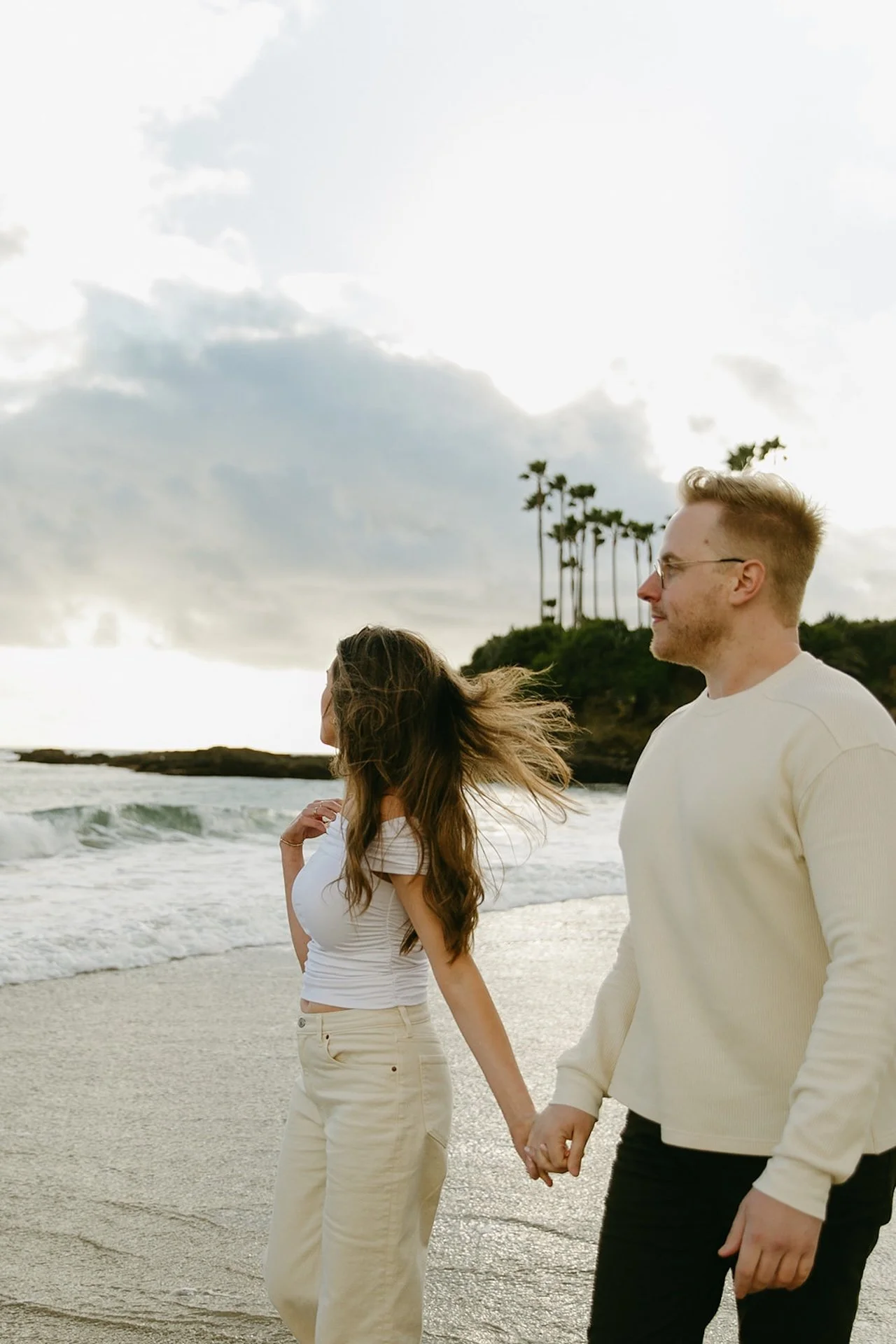 The engaged couple standing ankle-deep in the water, laughing together as the tide moves in along the sandy shoreline.