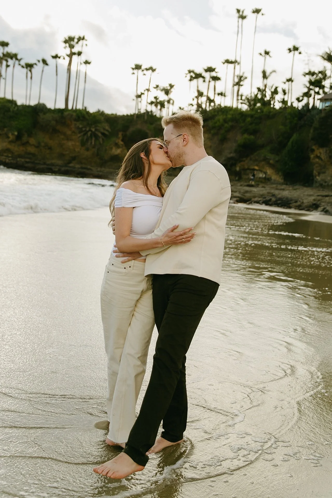 Soft coastal portrait of the engaged couple sharing a kiss near the water with palm-lined cliffs in the background.