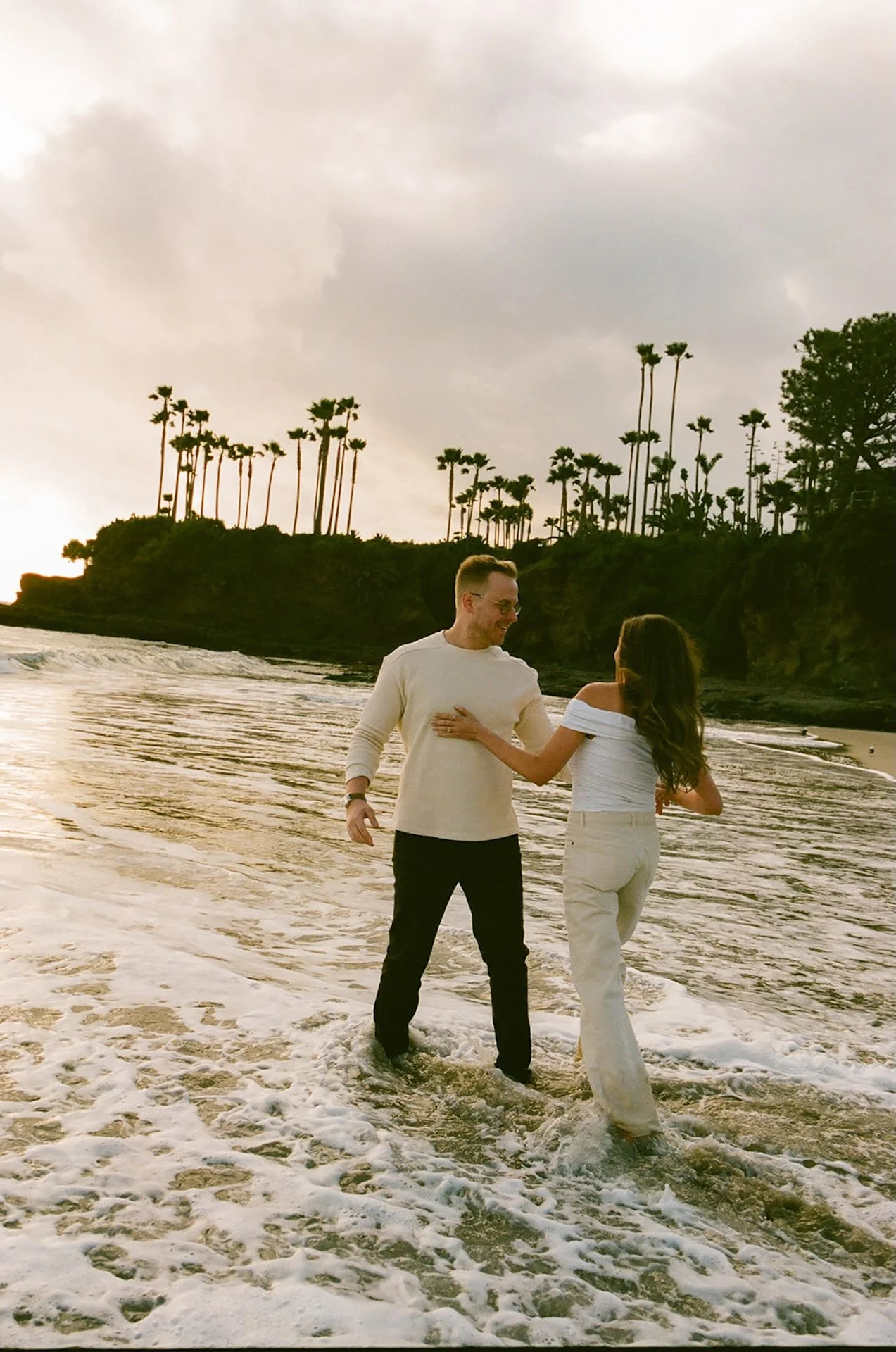 Wide shot of the couple splashing through shallow waves, laughing and reaching for each other during laguna beach engagement photos at shaws cove laguna beach.