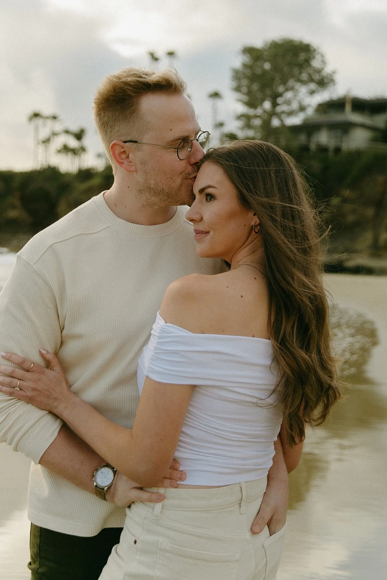 Close portrait of the engaged couple as he kisses her temple, soft coastal light and ocean water behind them.