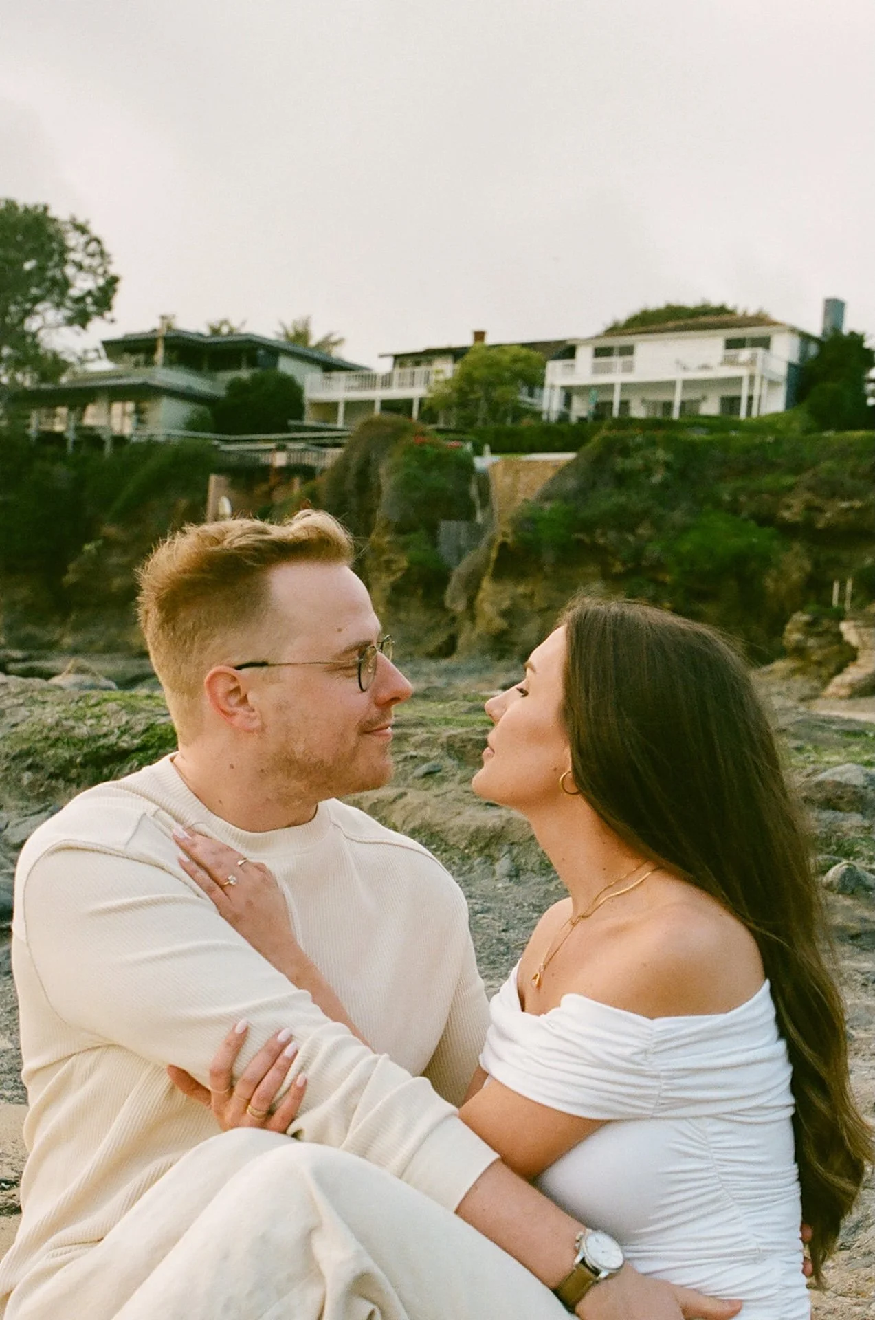 Seated portrait of the engaged couple on the sand, facing each other with rocky cliffs and coastal homes in the background.