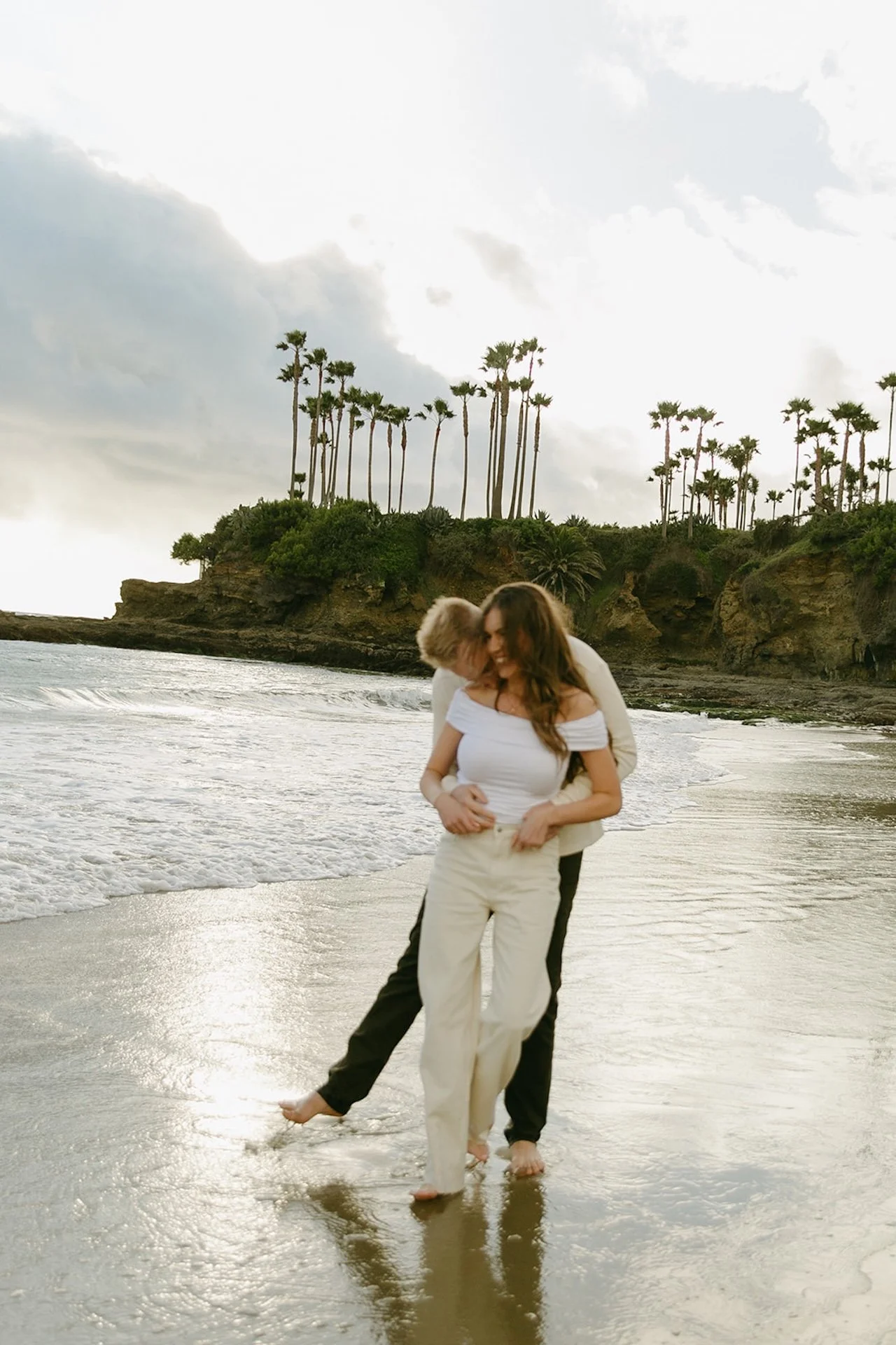 Playful candid of the engaged couple walking together through wet sand as waves reflect the cloudy coastal sky.