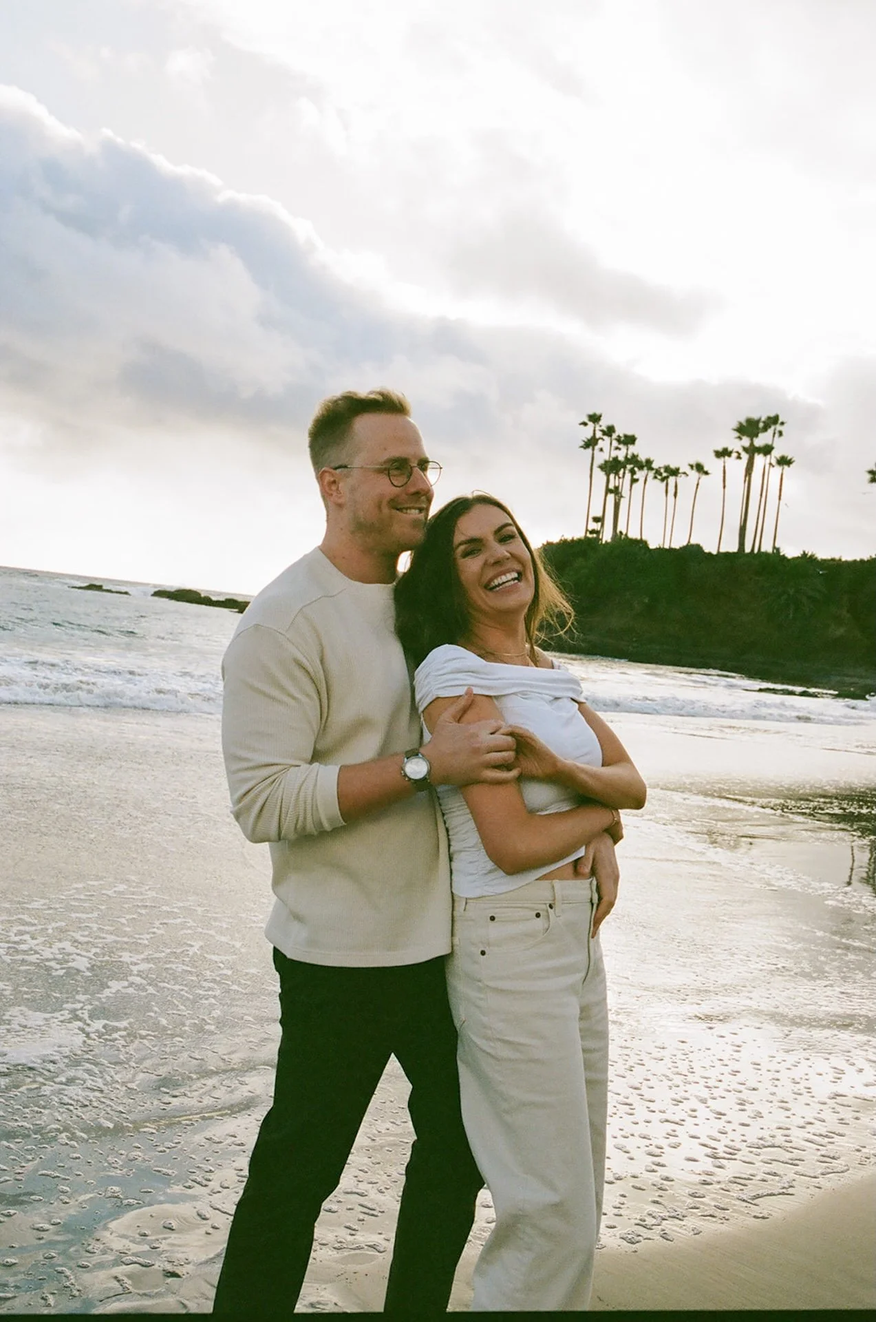 Wide shot of a couple embracing on a quiet beach with palm trees on the cliffs behind them, soft clouds and moody coastal light creating a cinematic engagement photo.