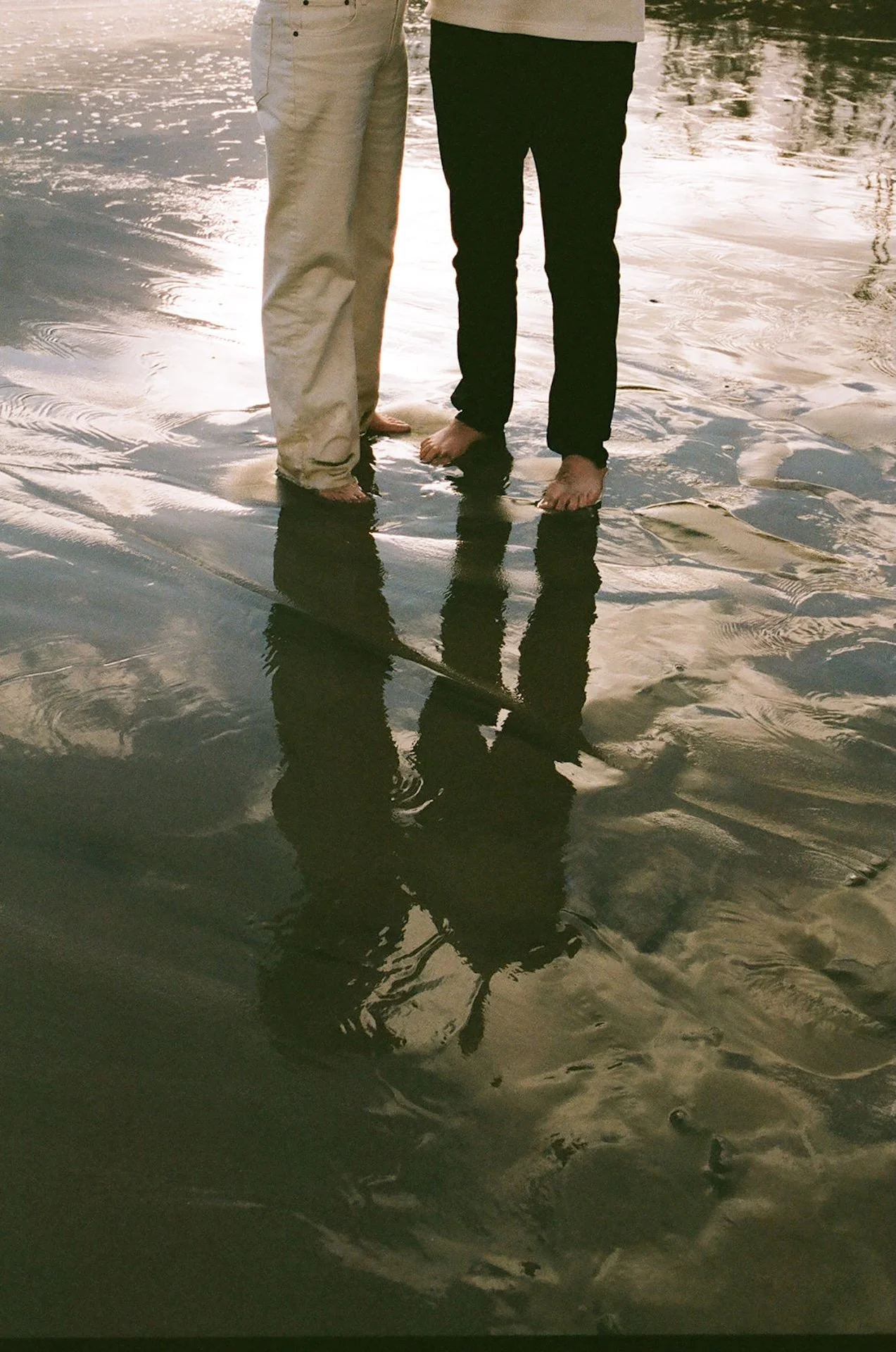Couple standing barefoot in shallow ocean water with reflections in the wet sand, wearing neutral outfits during a relaxed beach engagement photo session.