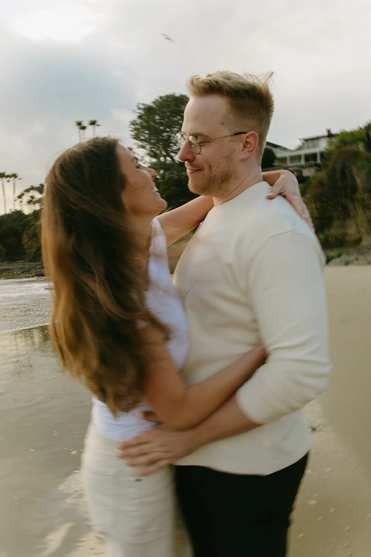 Close-up of a couple laughing while wrapped in each other’s arms near the shoreline, natural expressions and wind-blown hair during an intimate beach engagement session.