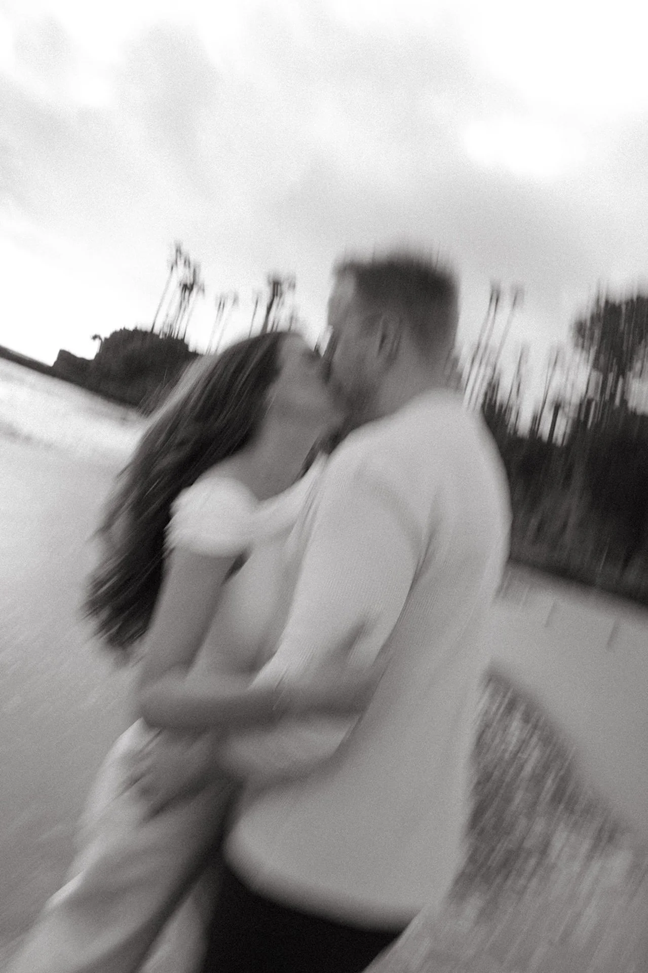 Black and white photo of a couple kissing with intentional motion blur, artistic and documentary-style engagement photo taken on the beach.