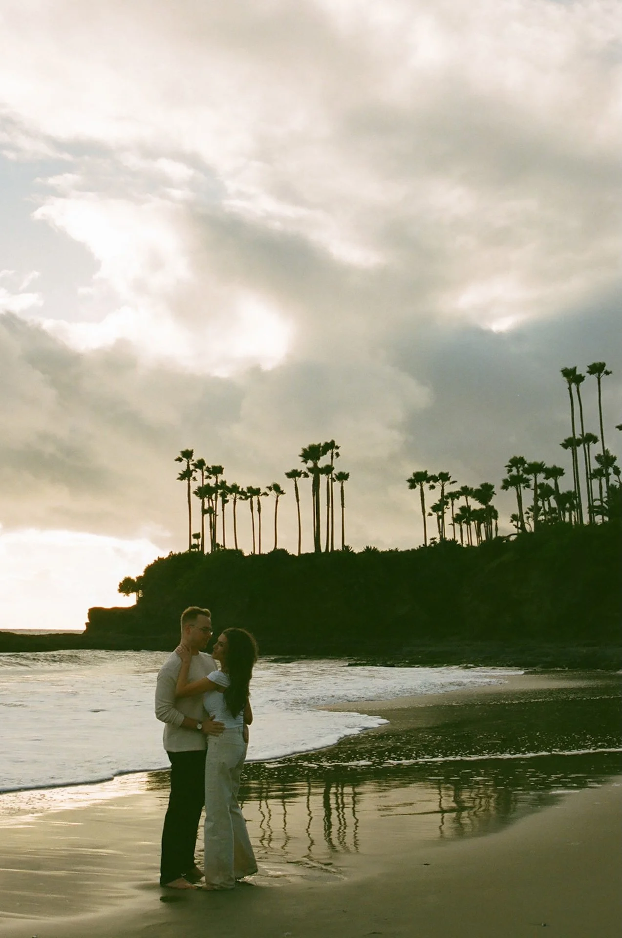 Wide shot of an engaged couple standing near the edge of the ocean, dramatic clouds and palm-lined cliffs framing a relaxed coastal engagement session.