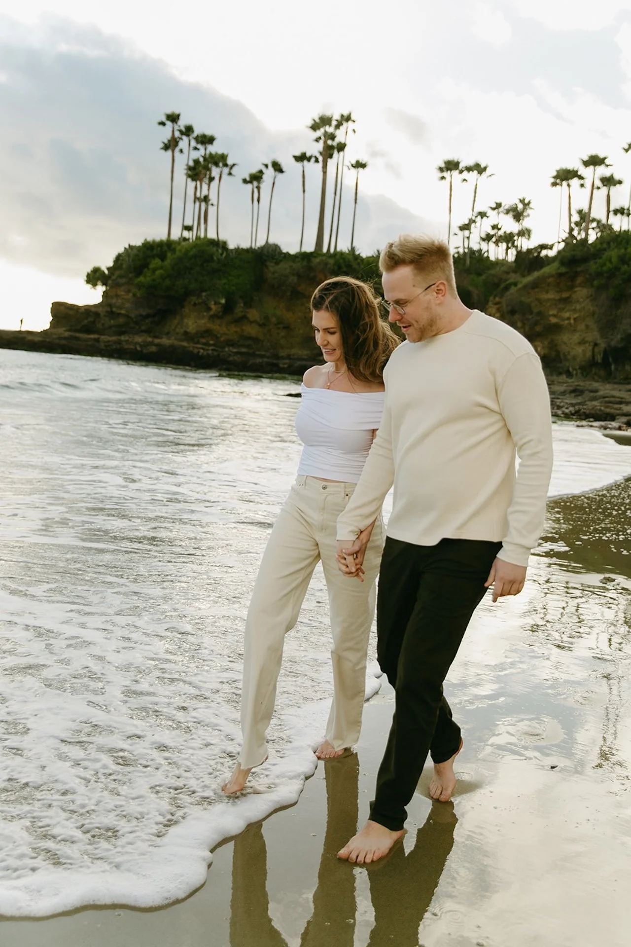 A couple holding hands while walking barefoot through shallow ocean water, dressed in neutral outfits with palm trees and coastal cliffs behind them.