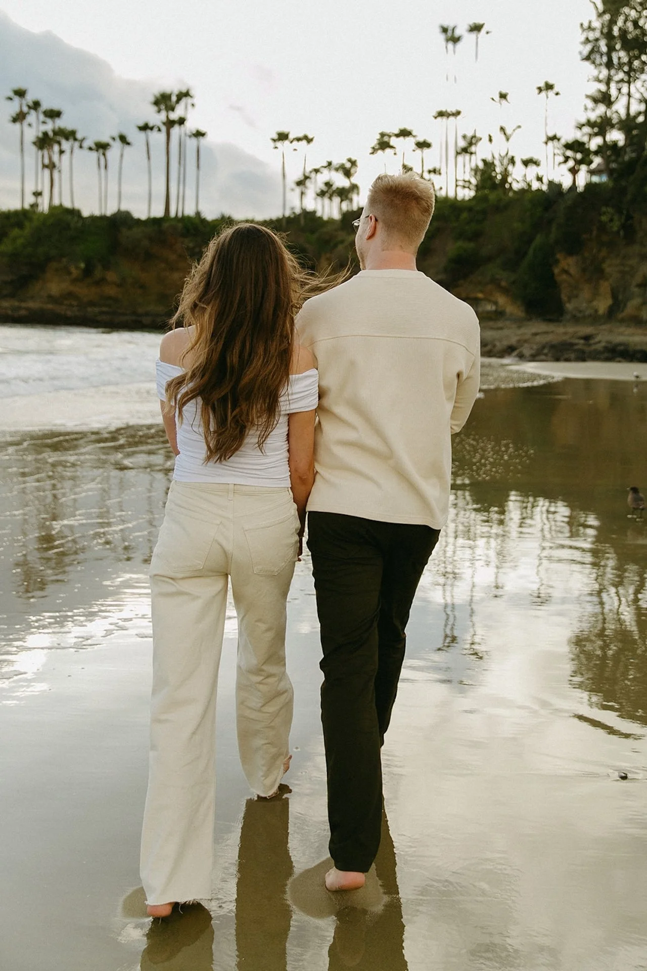 A couple standing side by side from behind, looking out toward the ocean with palm trees and coastal cliffs reflected in the wet sand.